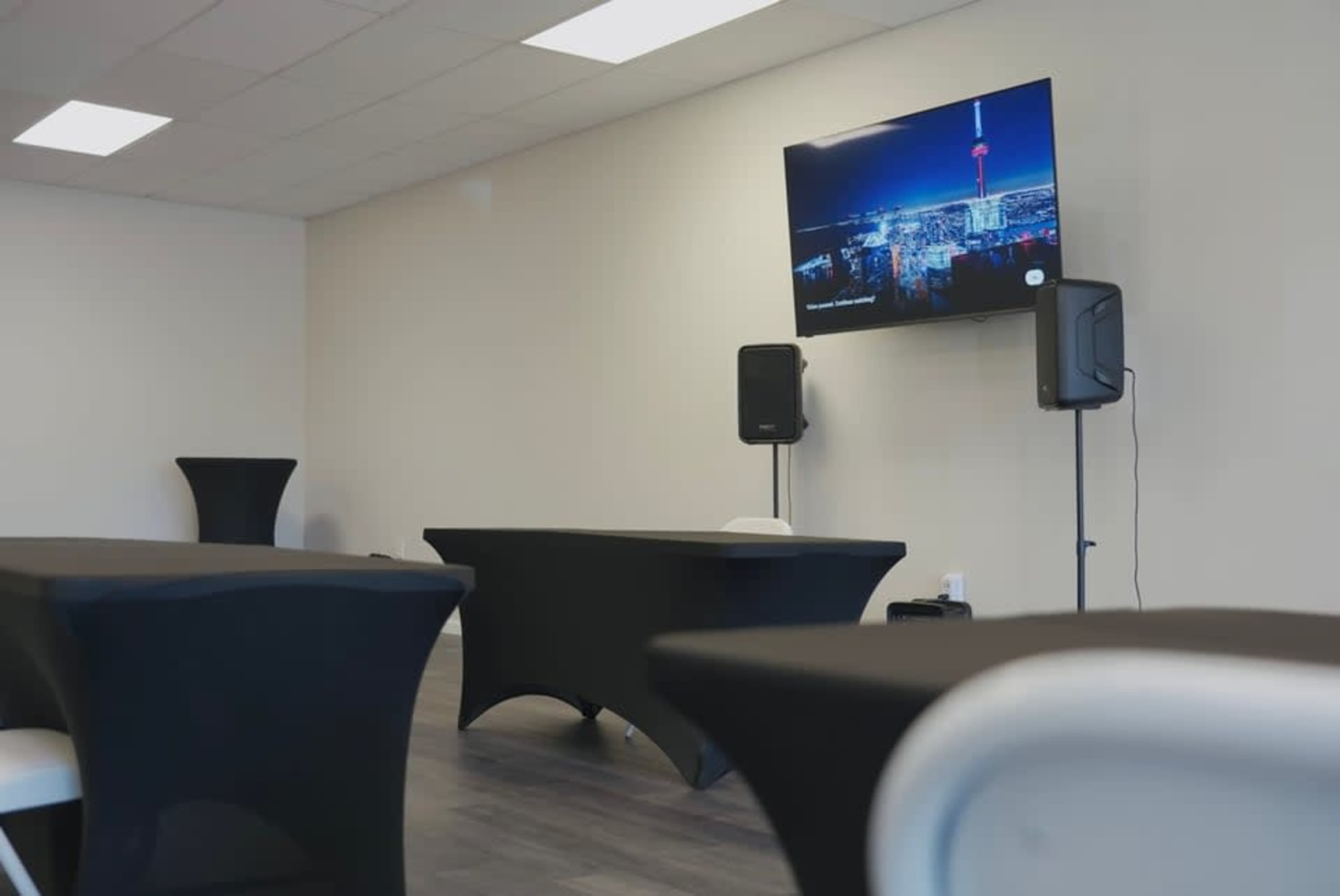 A spacious, minimally furnished event room features black tables and a large television displaying a cityscape, with audio equipment nearby.