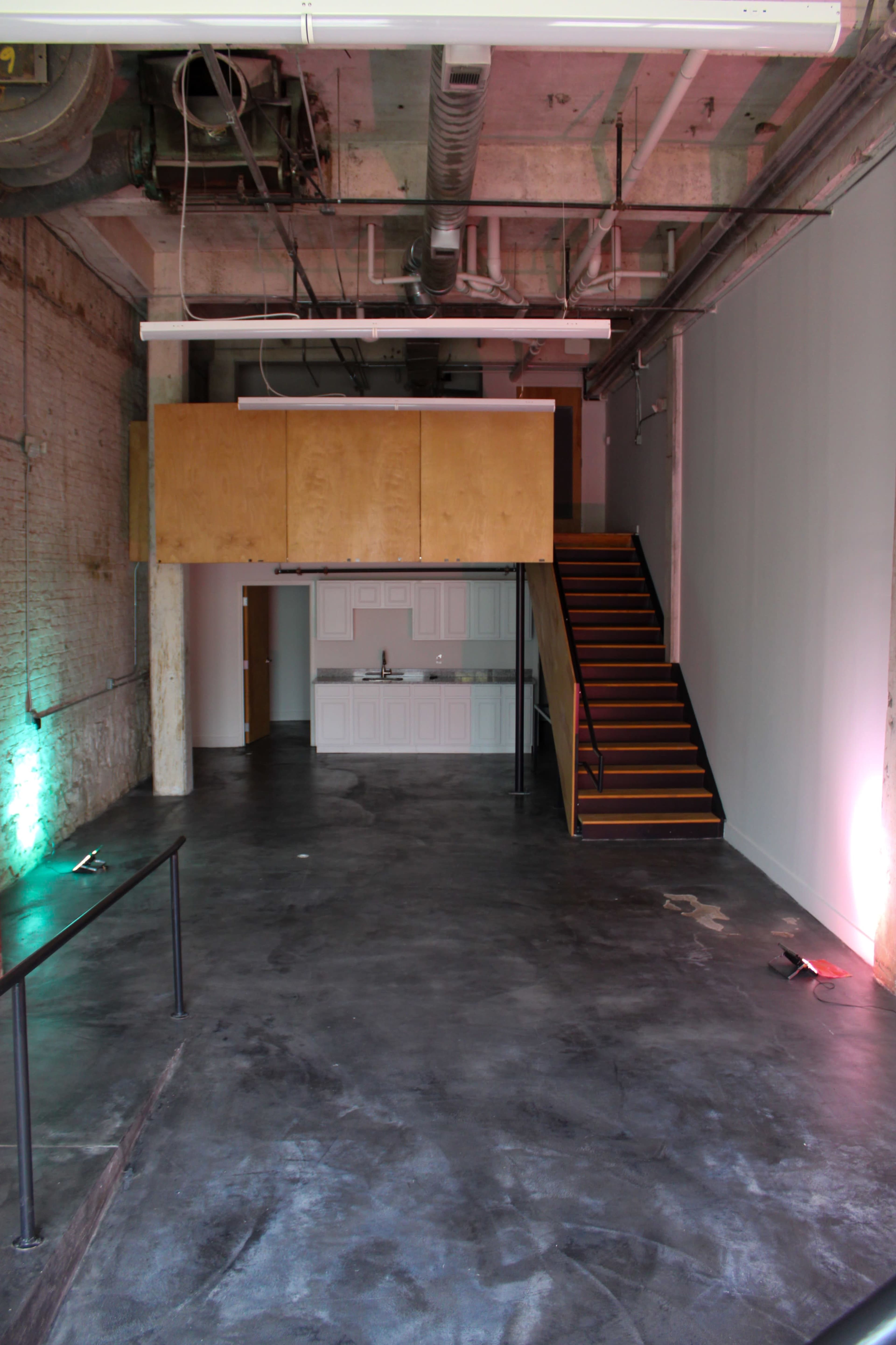 The image shows an industrial-style interior space featuring a concrete floor, exposed brick walls, a kitchenette area, and a staircase leading to an upper level.