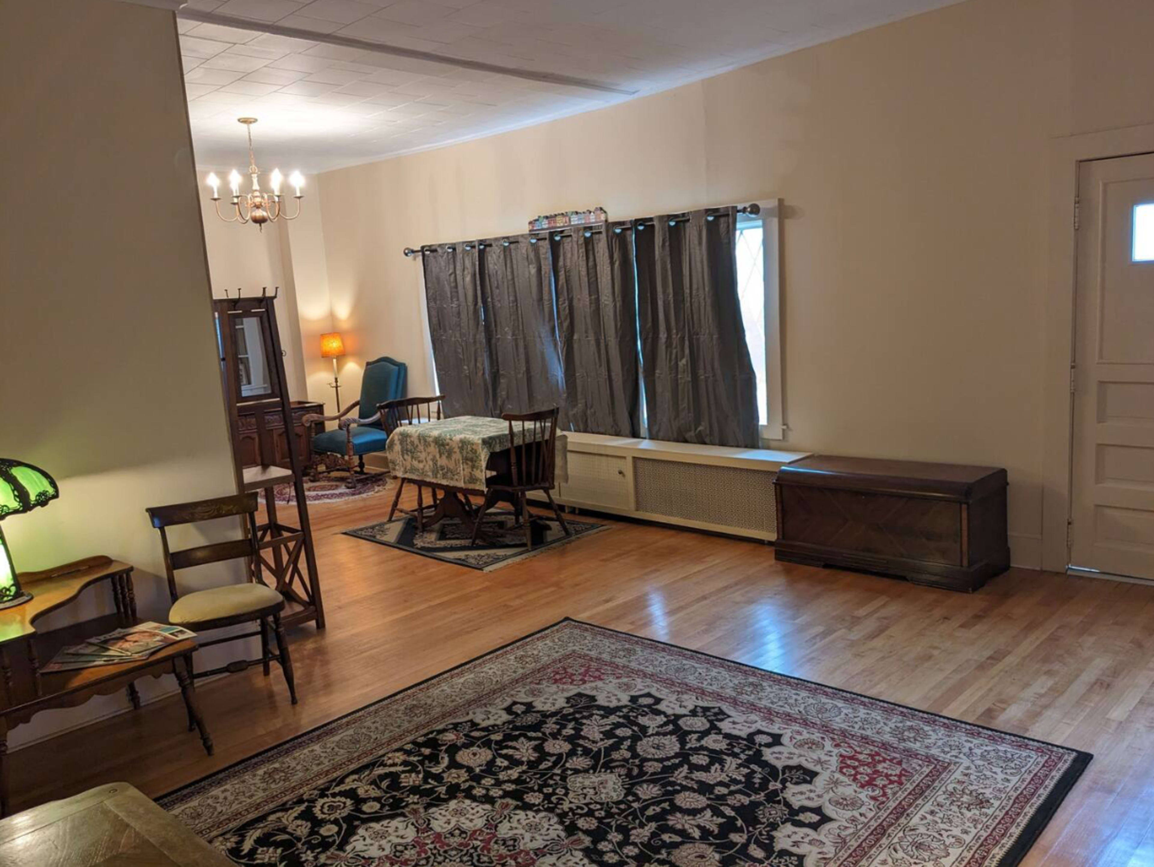 The image shows a spacious living room with wooden flooring, a central area rug, a dining table with chairs, and large curtains over a window.