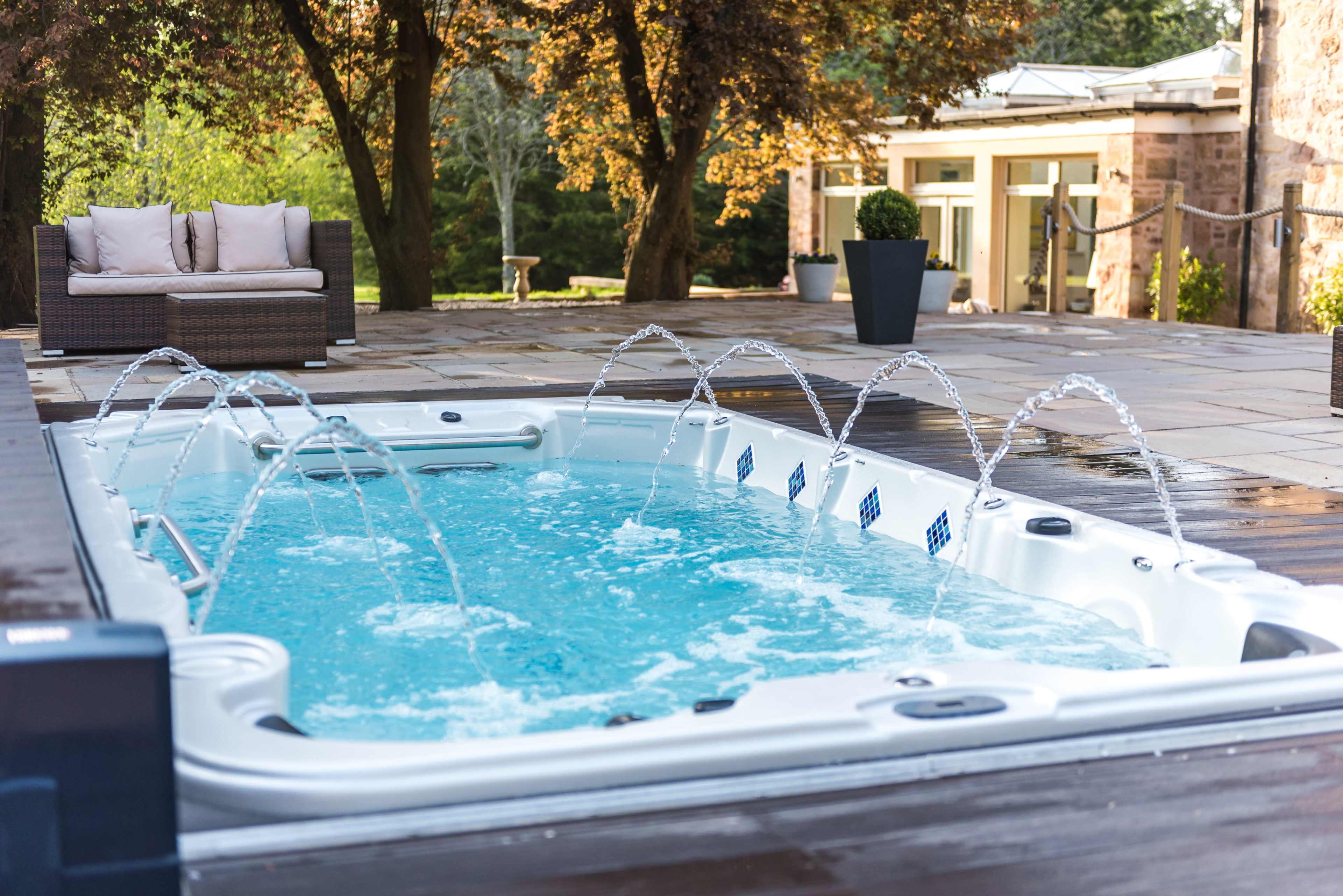 A hot tub with water jets is located on a wooden deck surrounded by trees and lounge furniture.