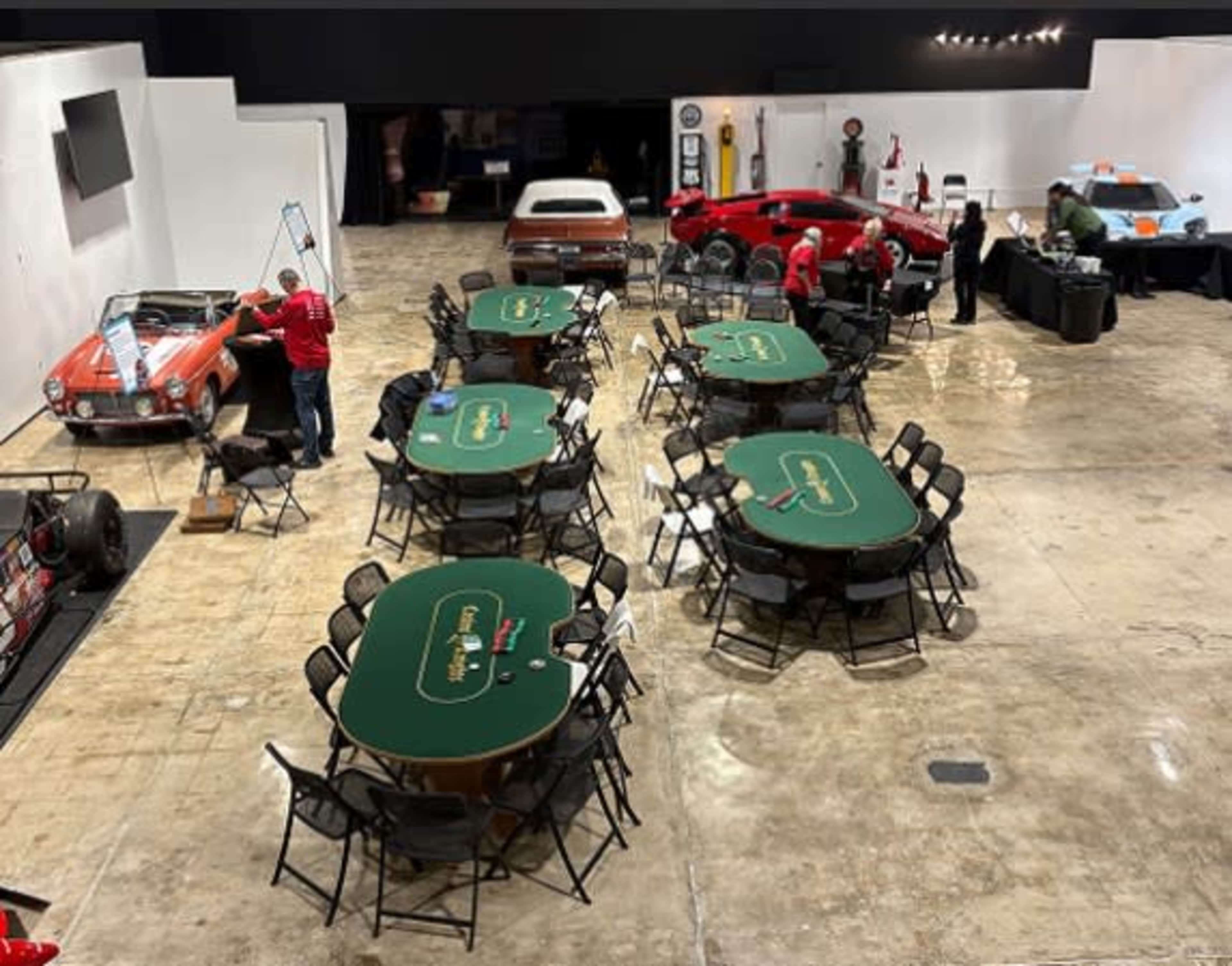 The image shows a spacious room arranged with several poker tables and classic cars displayed along the walls.