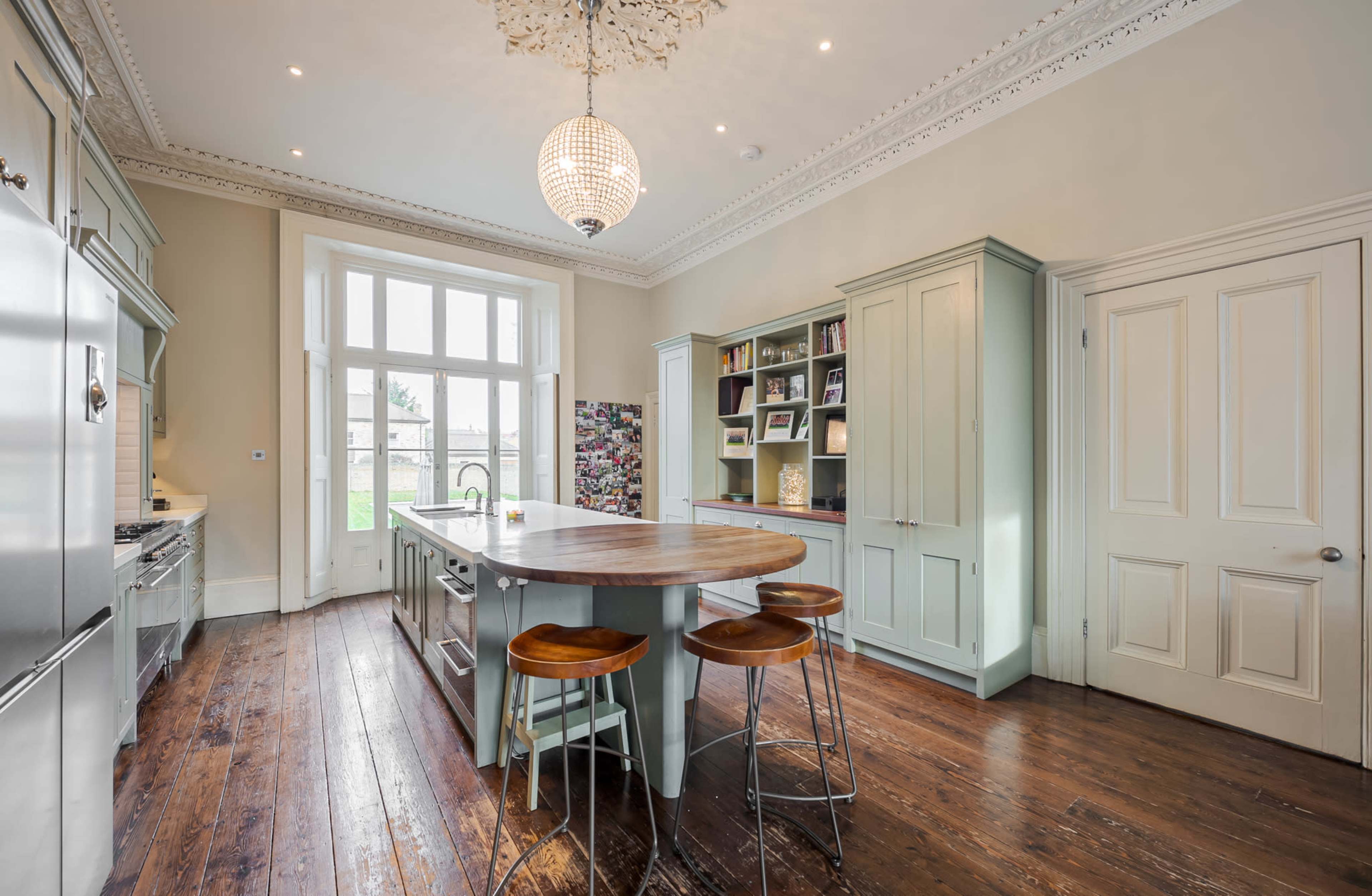The kitchen features a large wooden island with three stools, light green cabinetry, and large windows allowing natural light to fill the space.