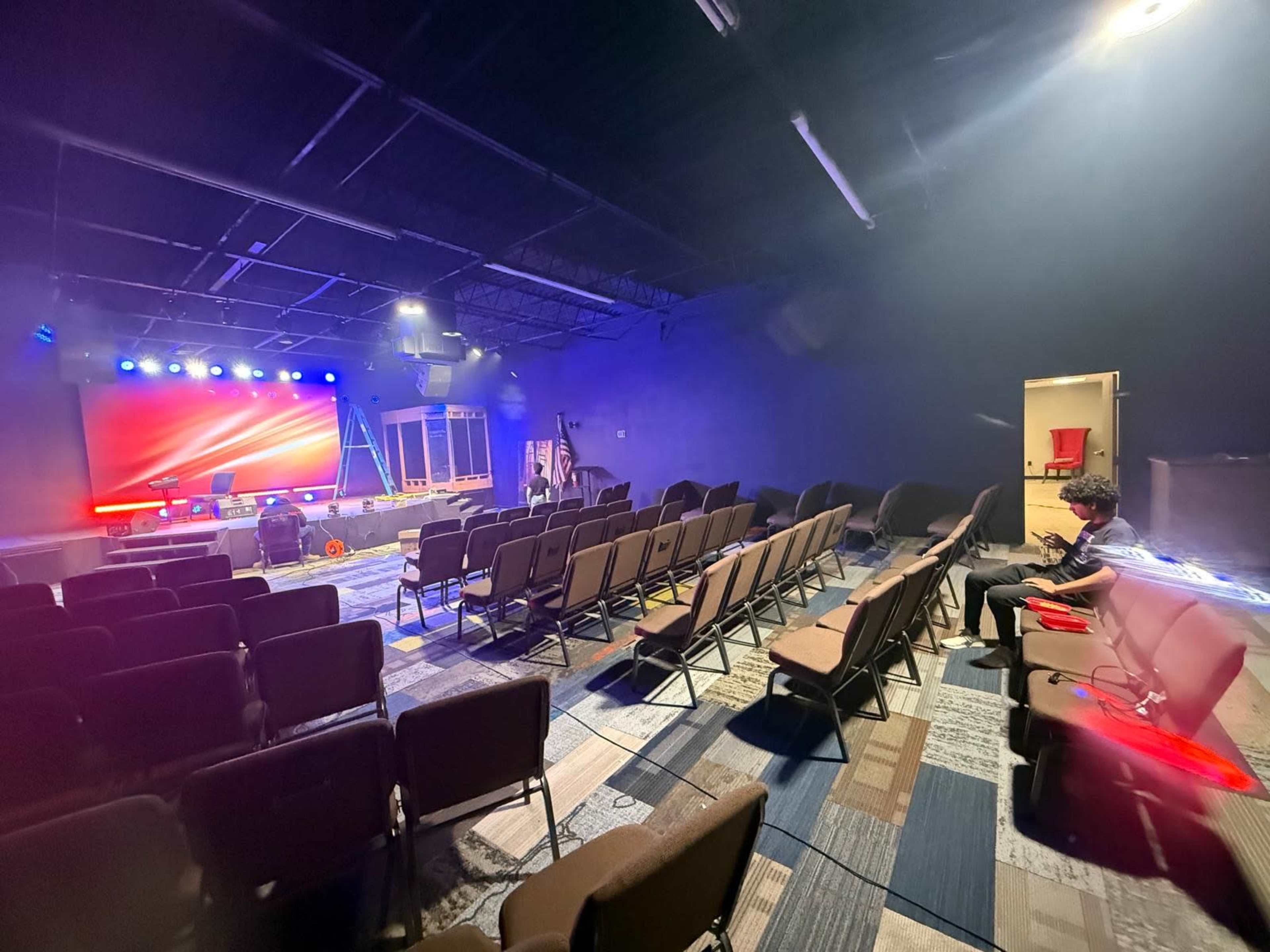 The image shows a dimly lit auditorium with rows of chairs facing a stage equipped with colorful lights and sound equipment, while a person sits at the end of one row.