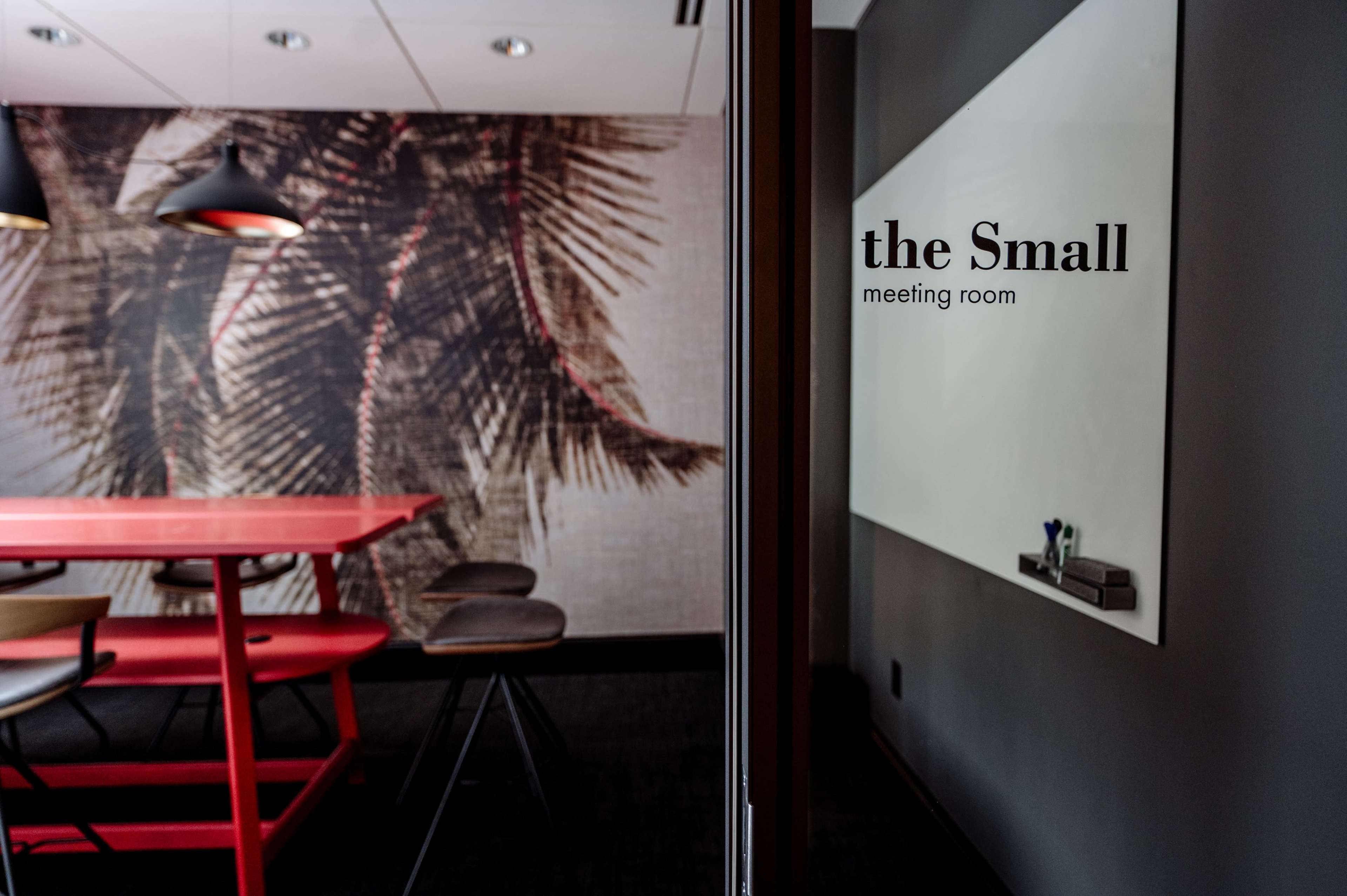 The image shows a modern meeting room with a red table, black stools, and a large wall mural featuring palm leaves.