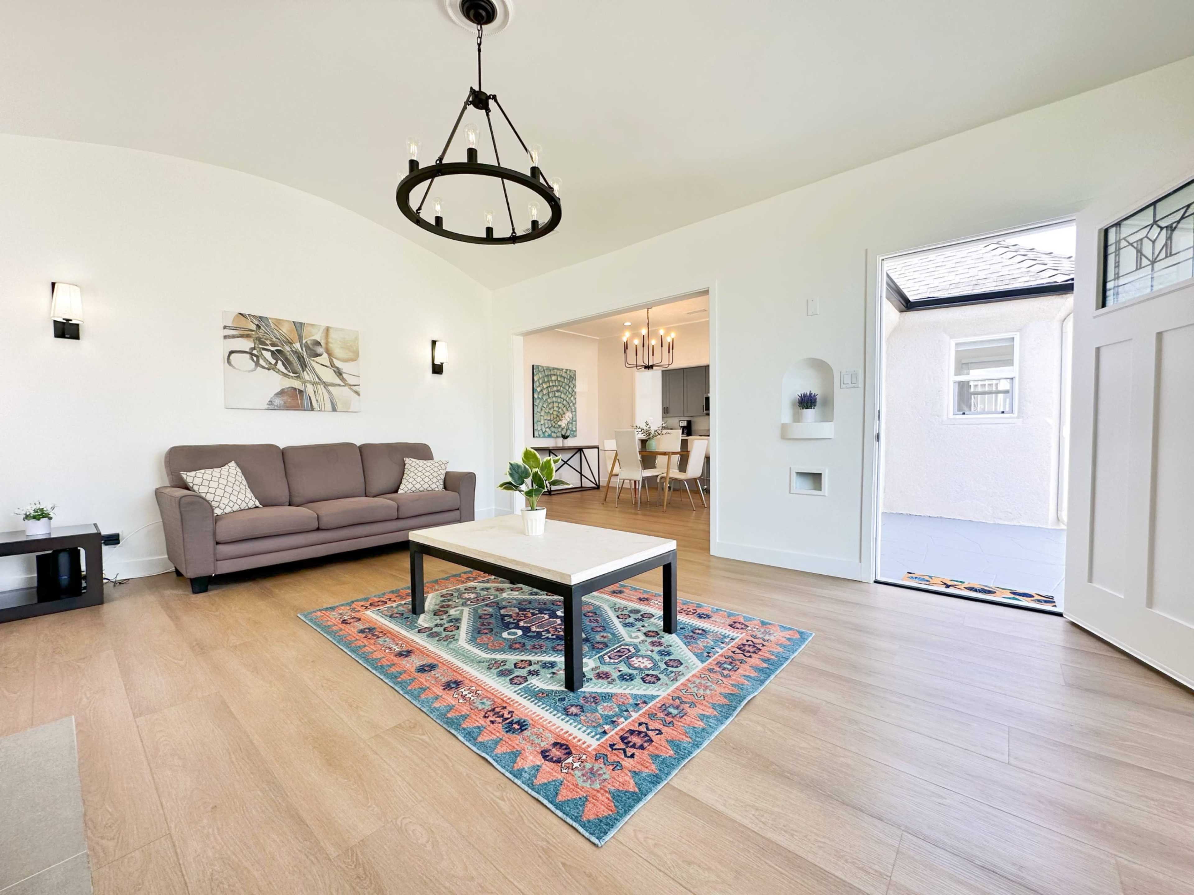 The image shows a bright living room with a chandelier, a brown sofa, a coffee table on a colorful rug, and an open door leading to another room.