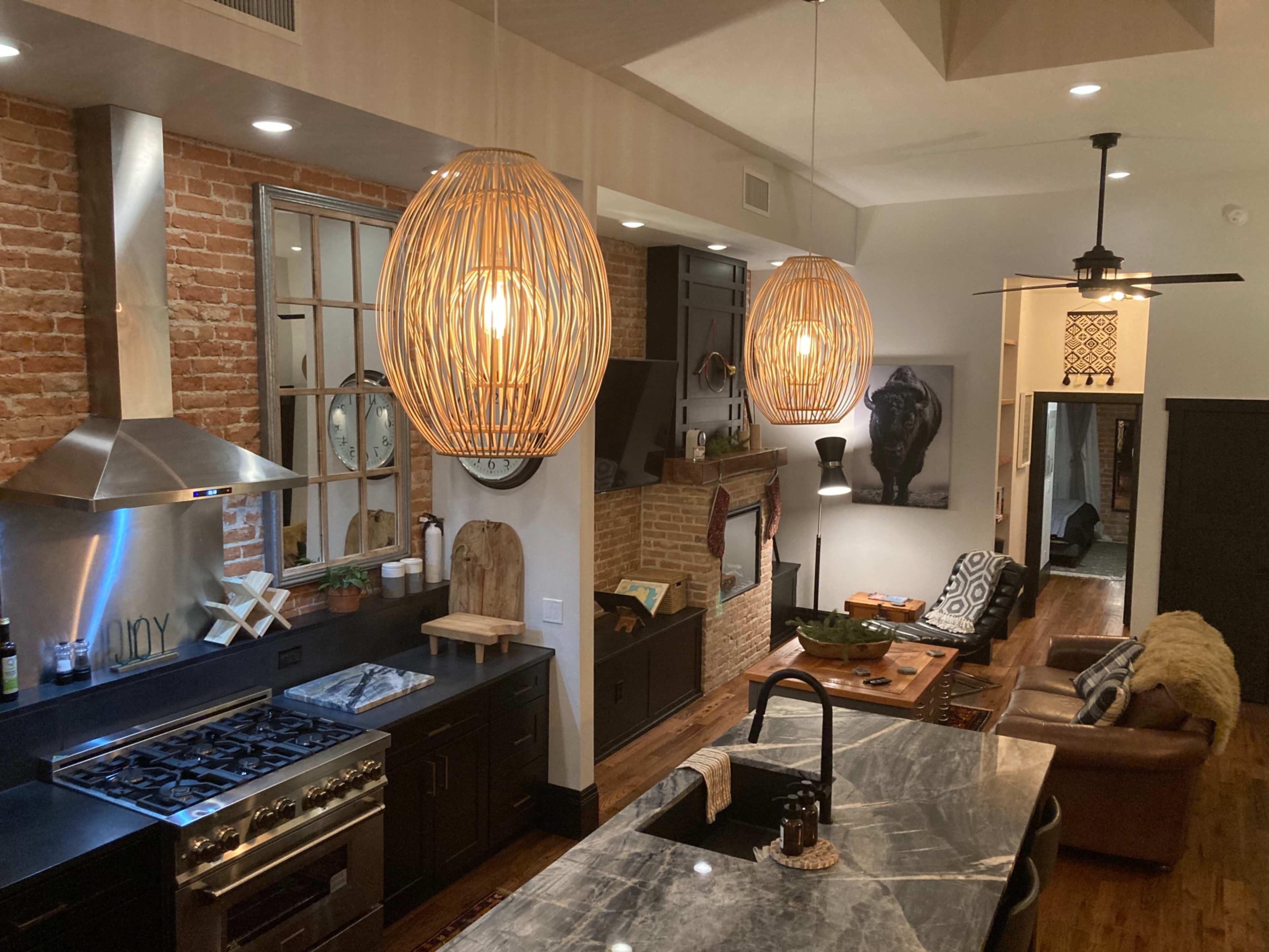 The image shows a modern kitchen with a central island, featuring a stainless steel stove and woven pendant lights, alongside a living area with leather seating and exposed brick walls.
