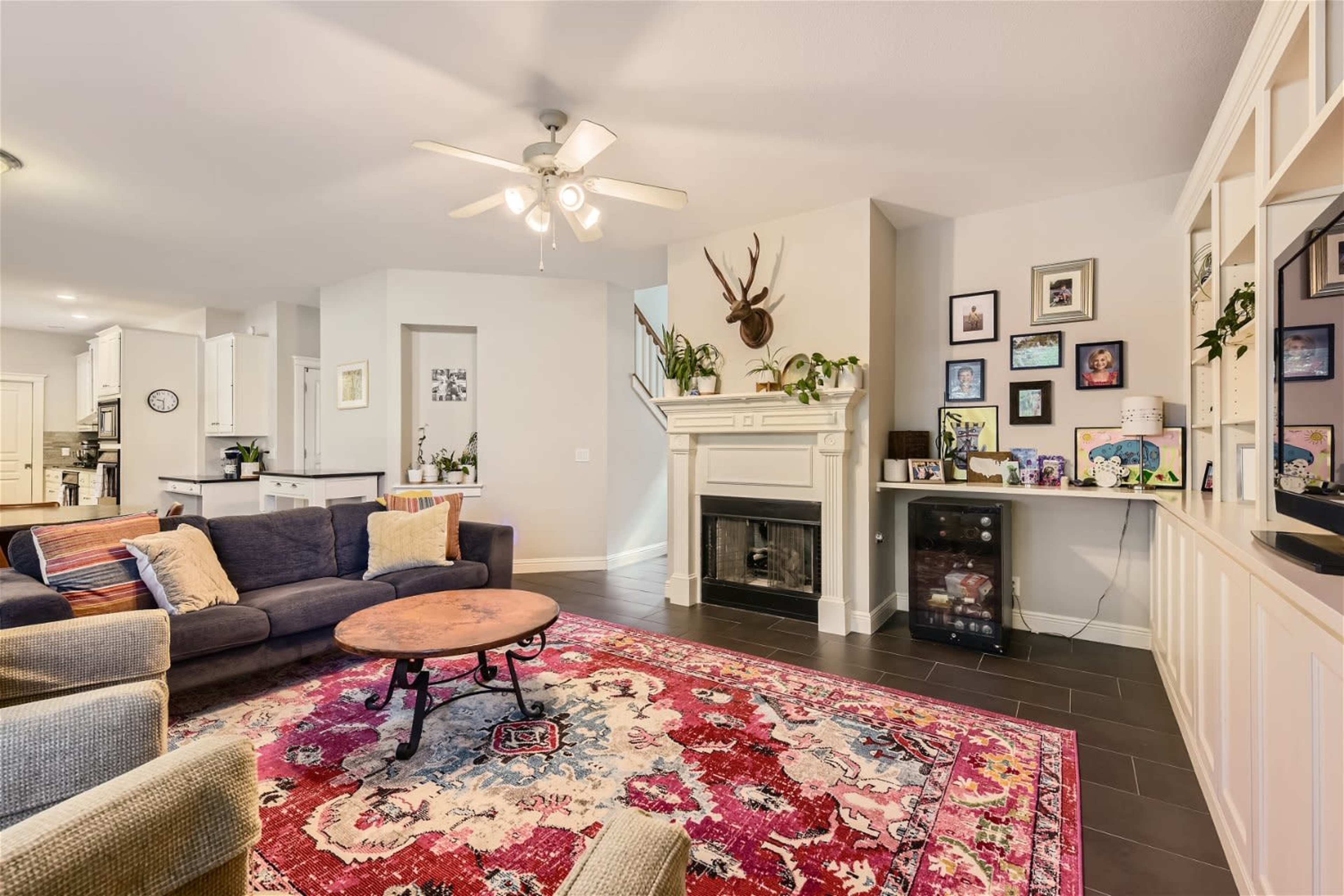 The living room features a patterned area rug, a circular coffee table, a fireplace with decorative items, and a wall displaying framed photos and art.