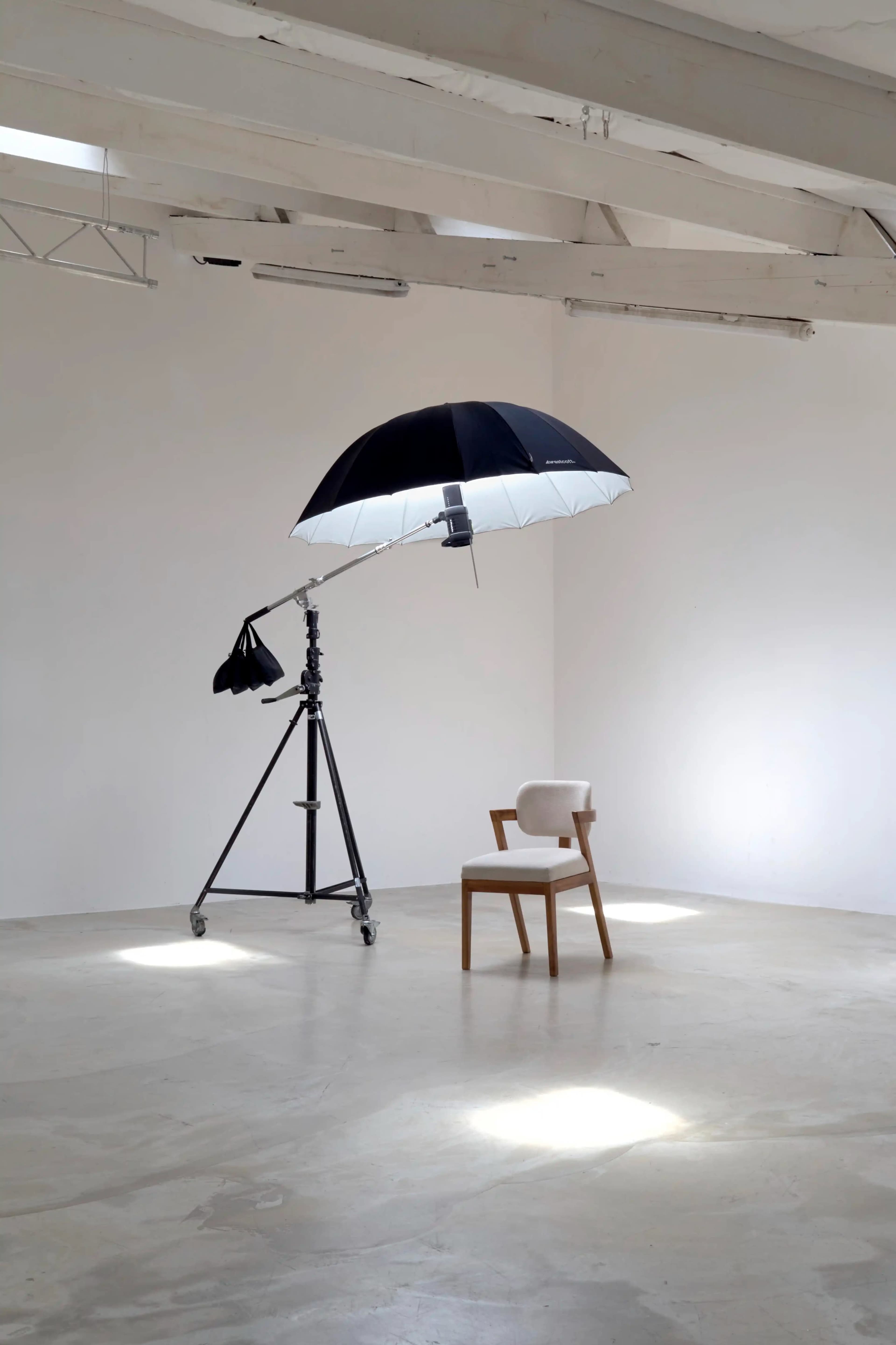 A black and white umbrella is mounted on a tripod above a wooden chair in a minimalist room with white walls and a concrete floor.