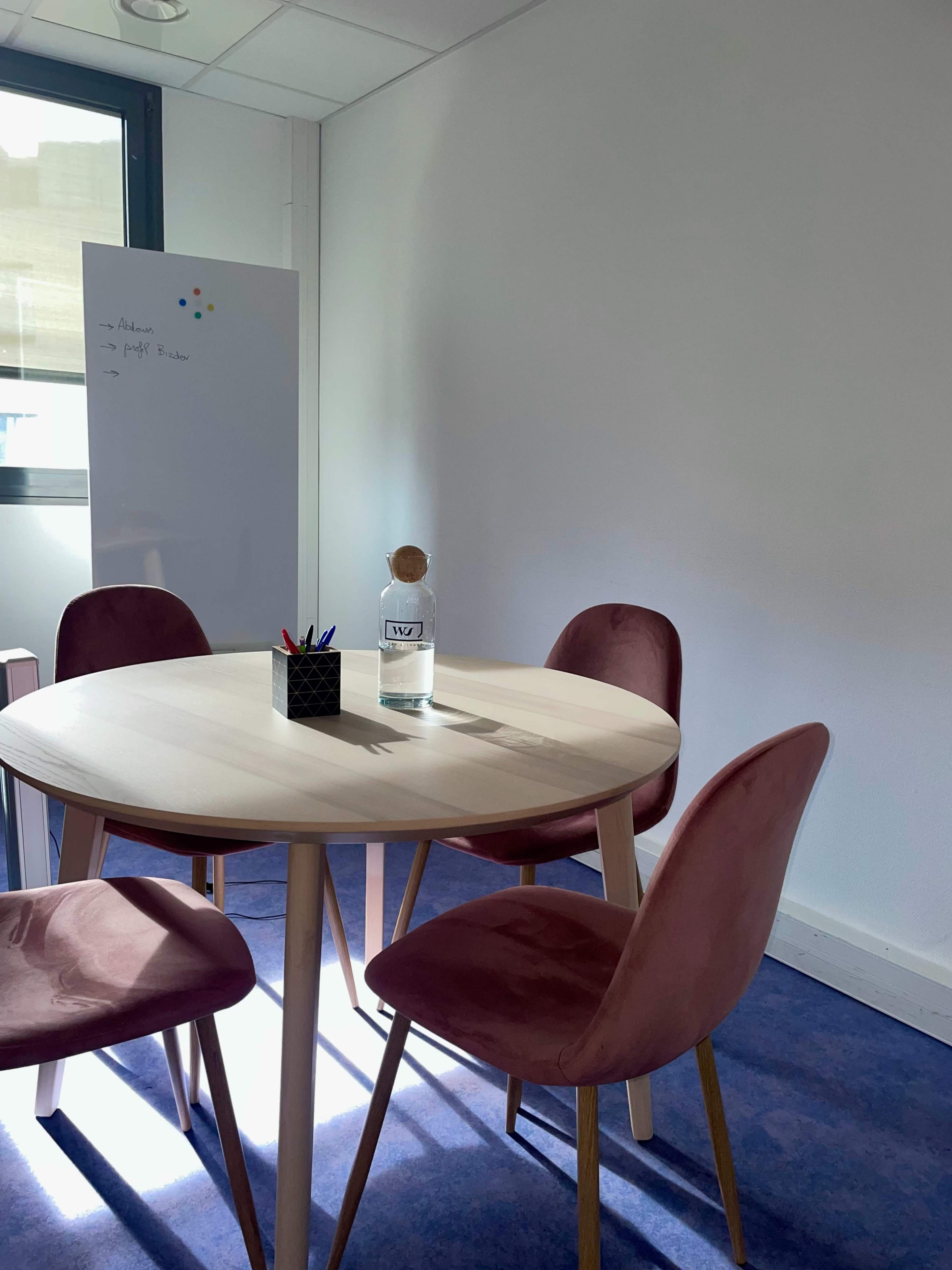 A small meeting room features a round wooden table surrounded by four pink chairs, with a glass water bottle and a pen holder on the table.