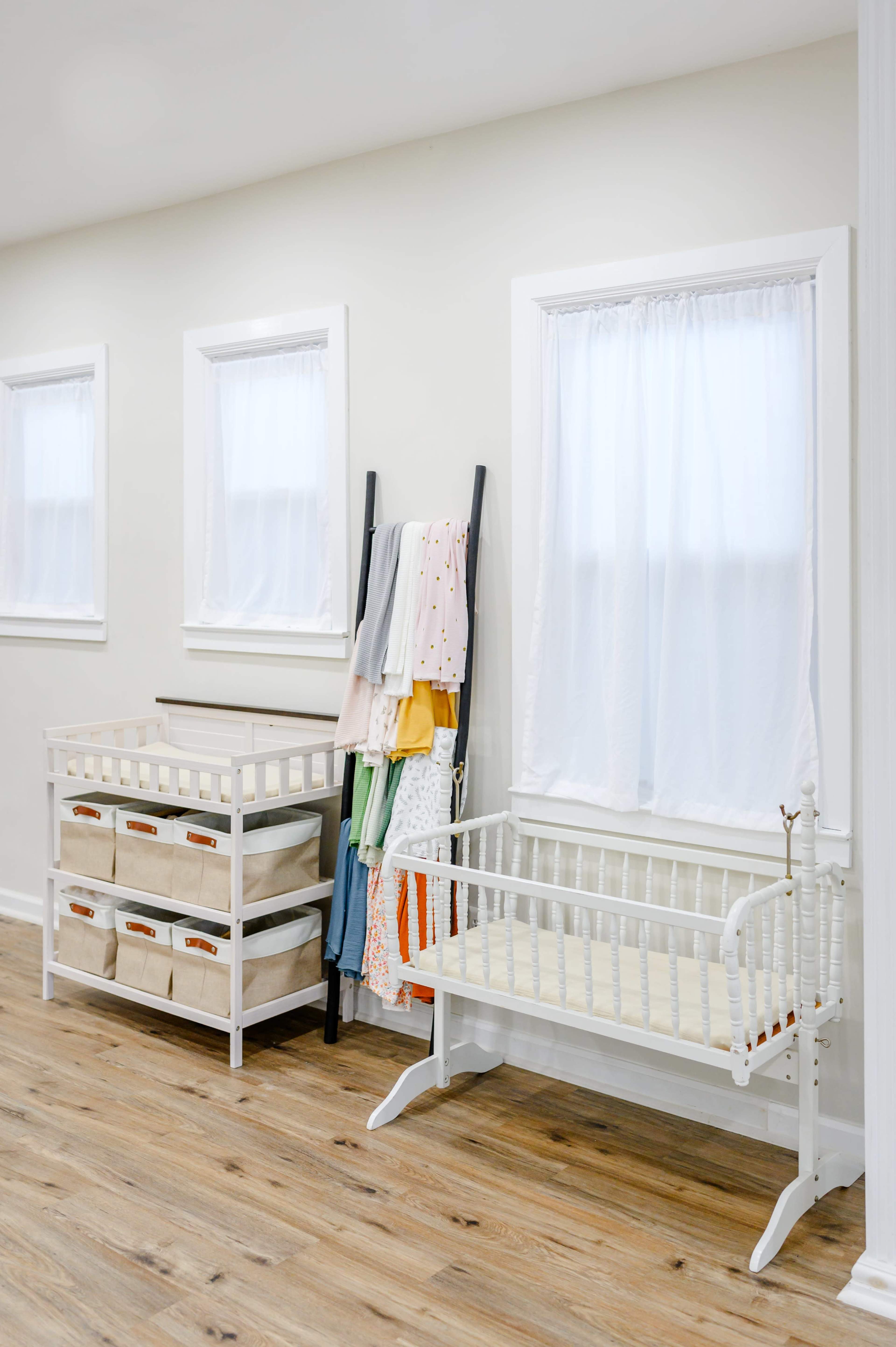 The image shows a bright room with two windows, a white changing table with storage baskets, and a white crib beside a rack holding colorful cloths.