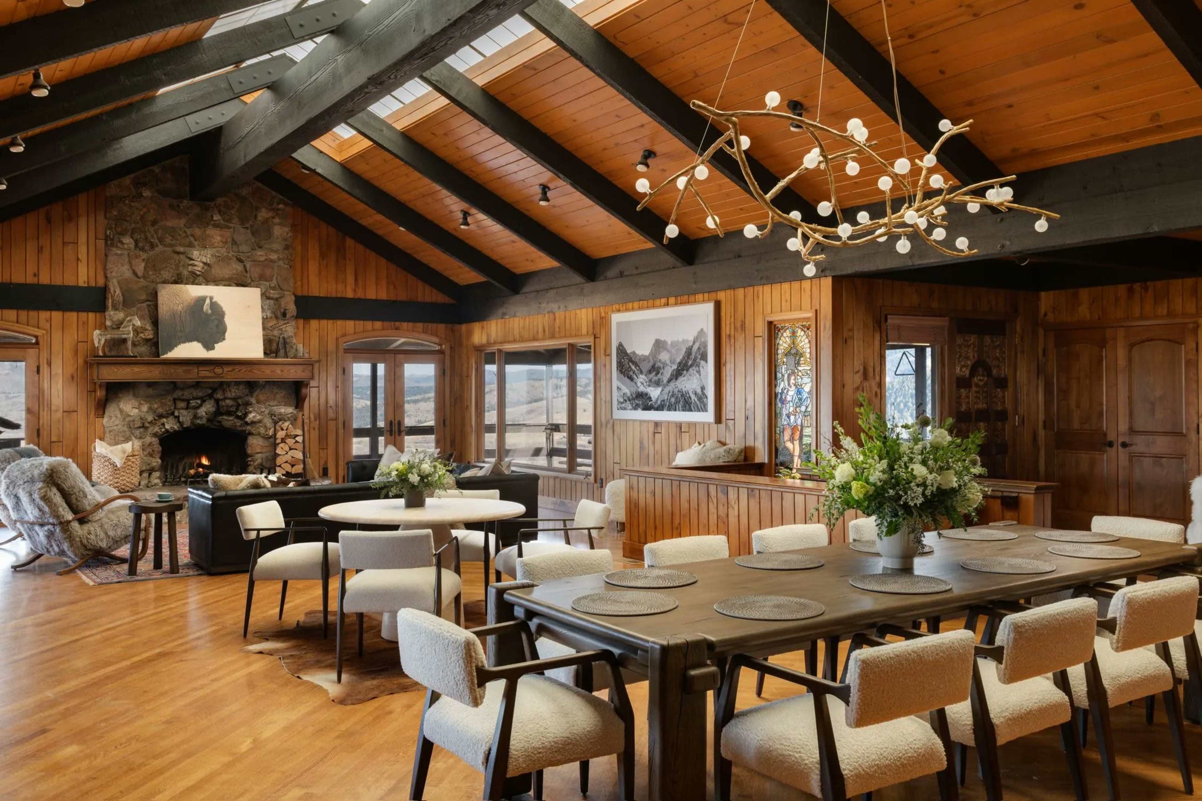 The image depicts a cozy dining area with wooden beams, a stone fireplace, and a large dining table surrounded by upholstered chairs.