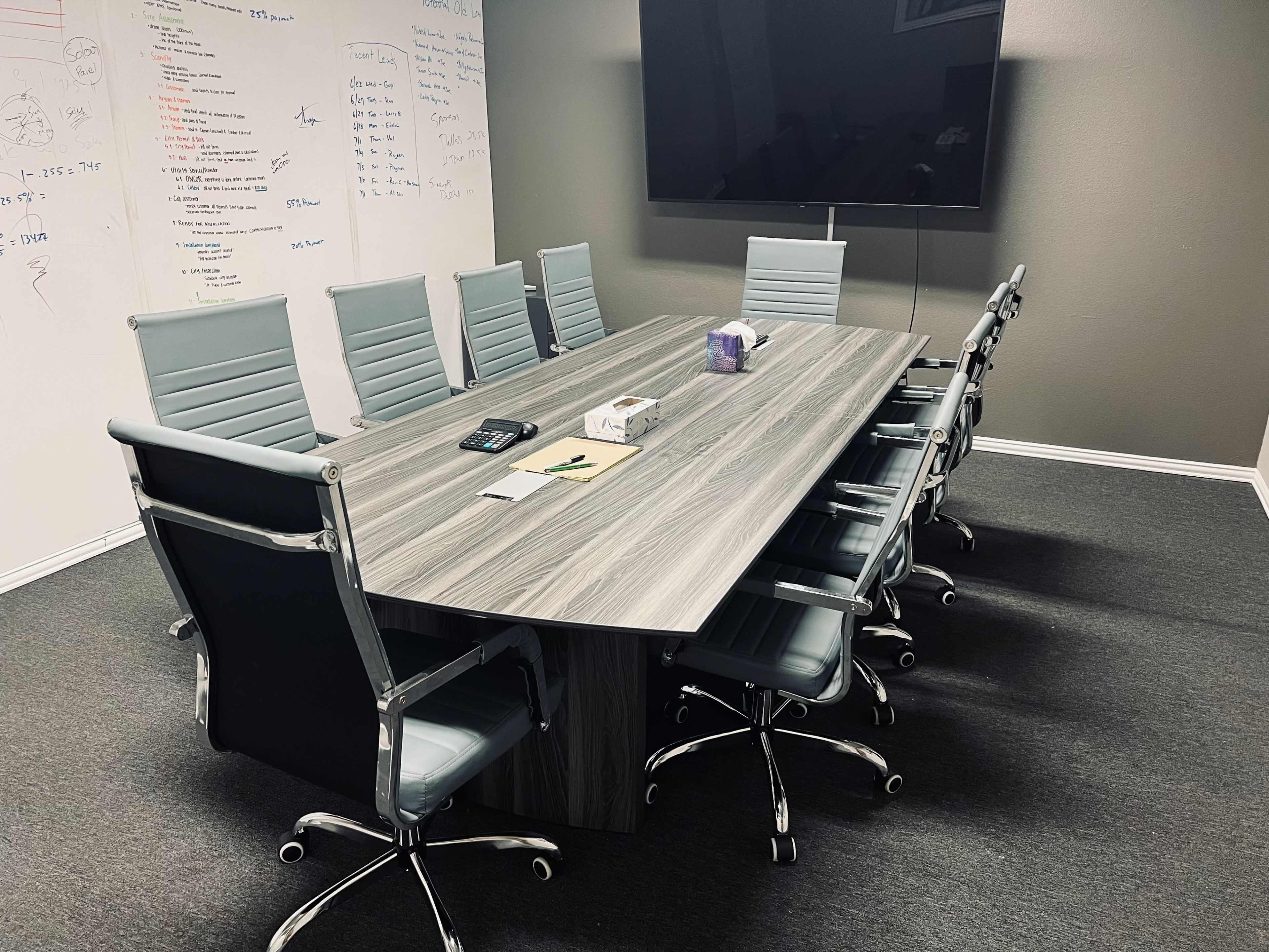 A modern conference room features a large table surrounded by multiple ergonomic chairs, with a wall-mounted screen and whiteboard covered in notes.