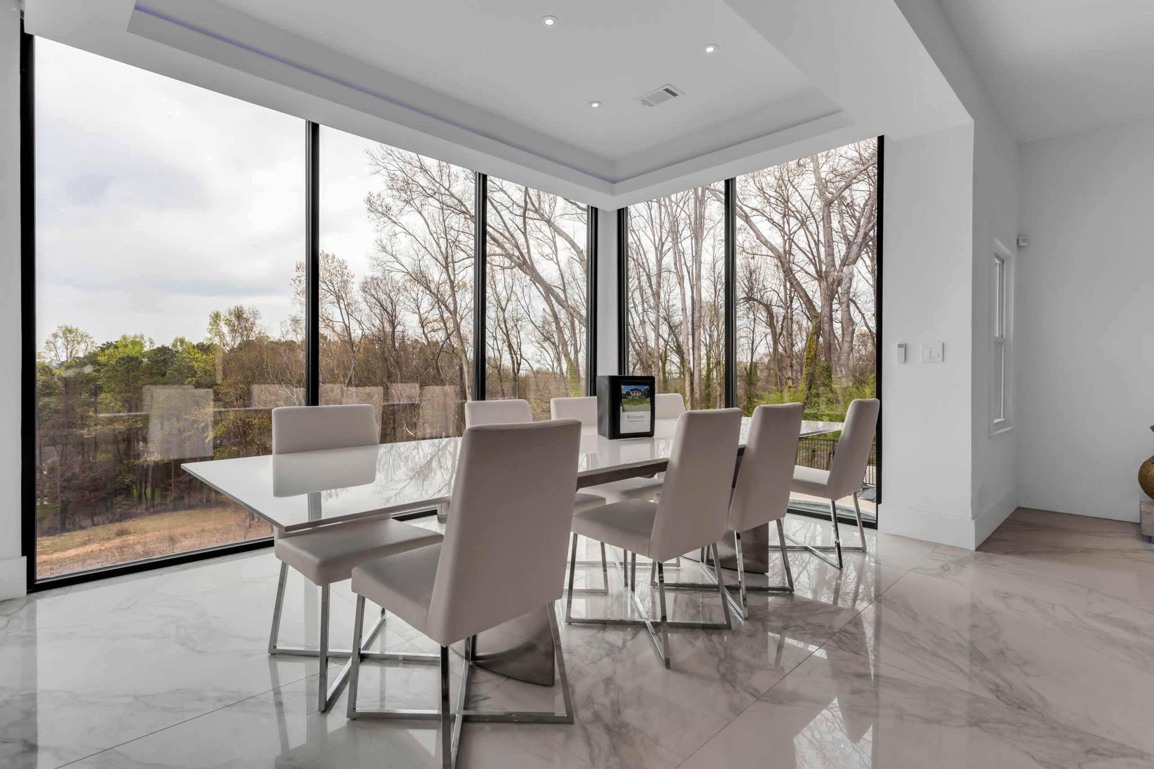 A modern dining room features a glass-topped table surrounded by white chairs, with large windows offering a view of a wooded area.