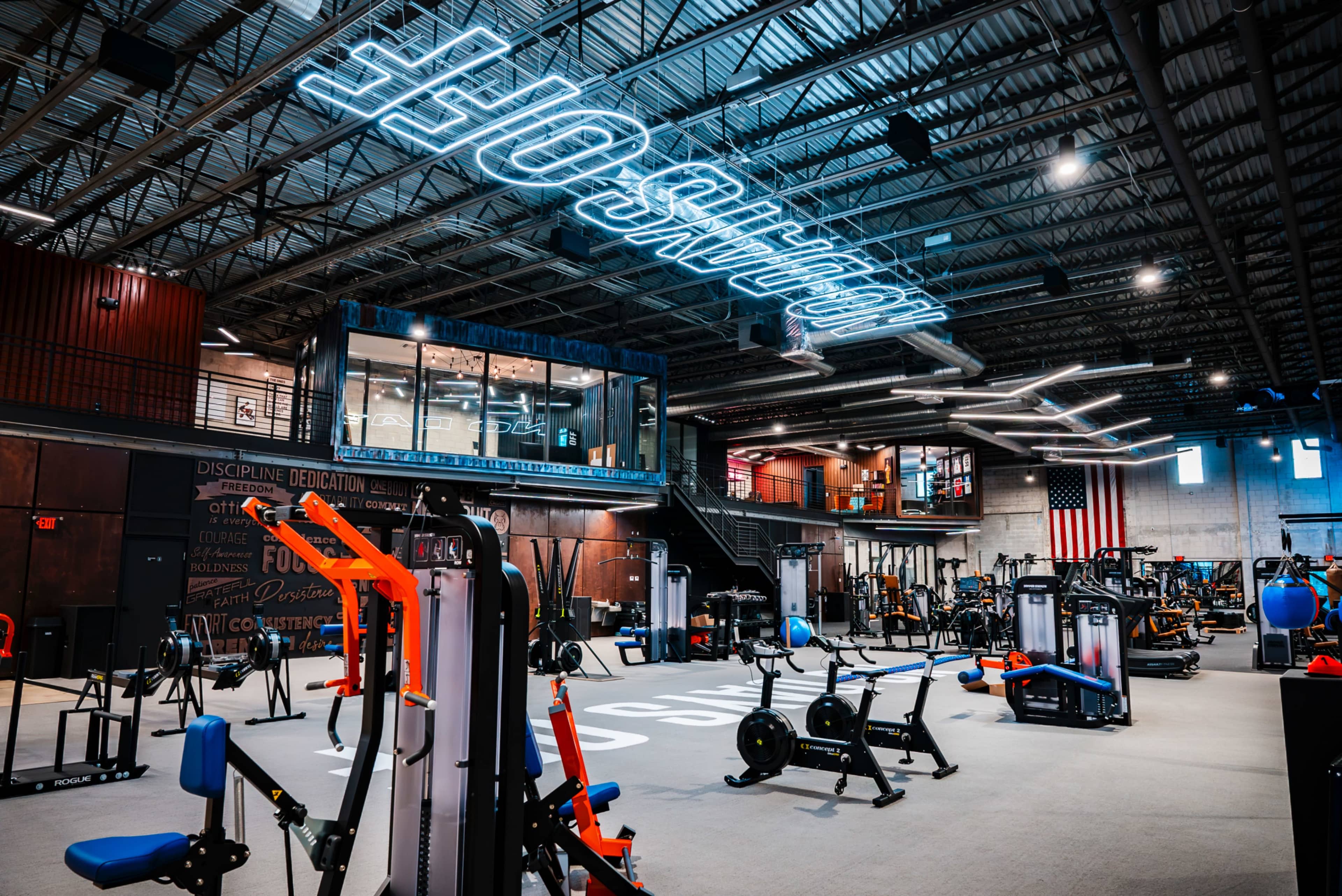 The image shows a spacious gym filled with various exercise equipment, including weight machines and free weights, under bright neon signage.