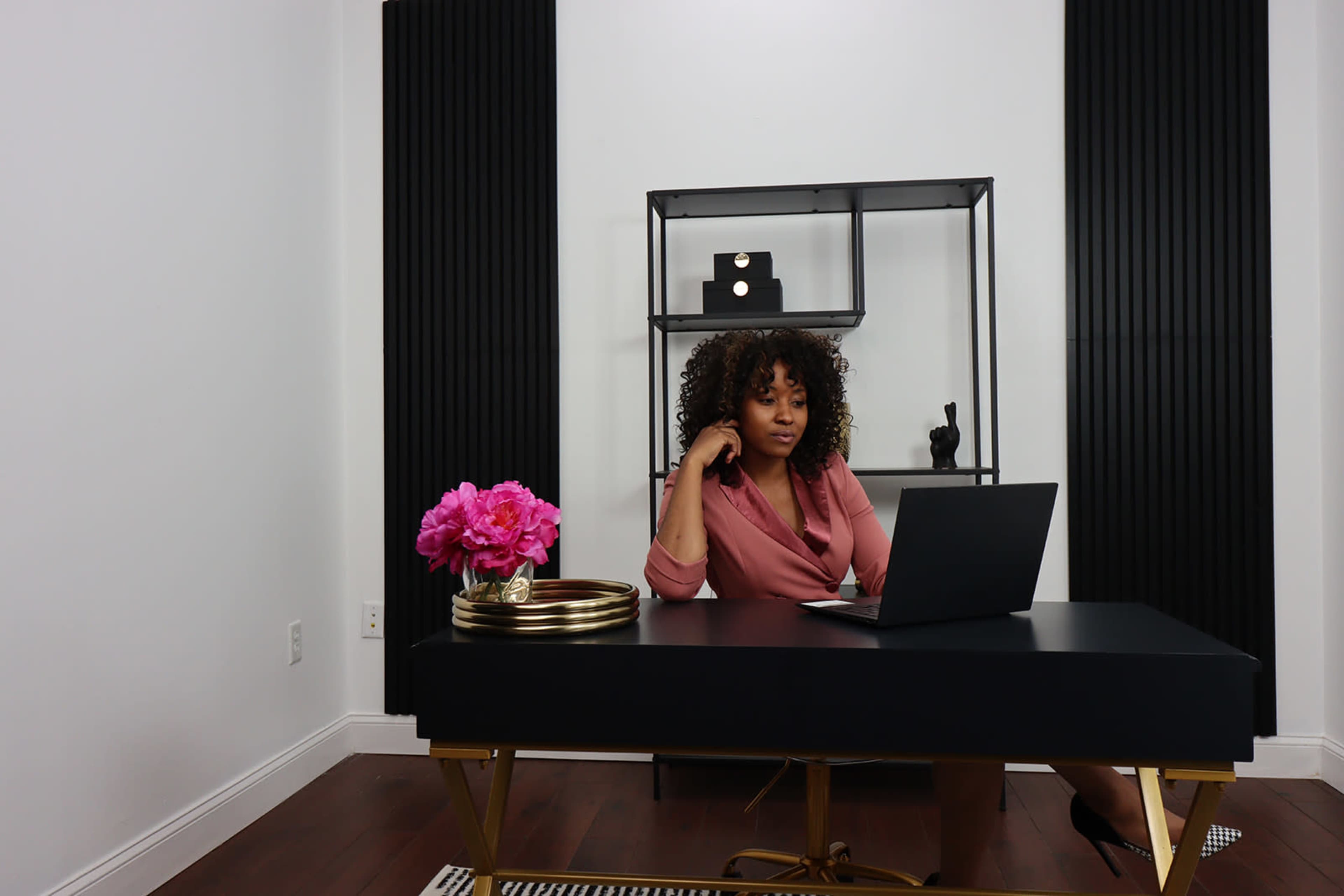A woman with curly hair sits at a black desk, working on a laptop in a modern office decorated with a floral arrangement and shelves.