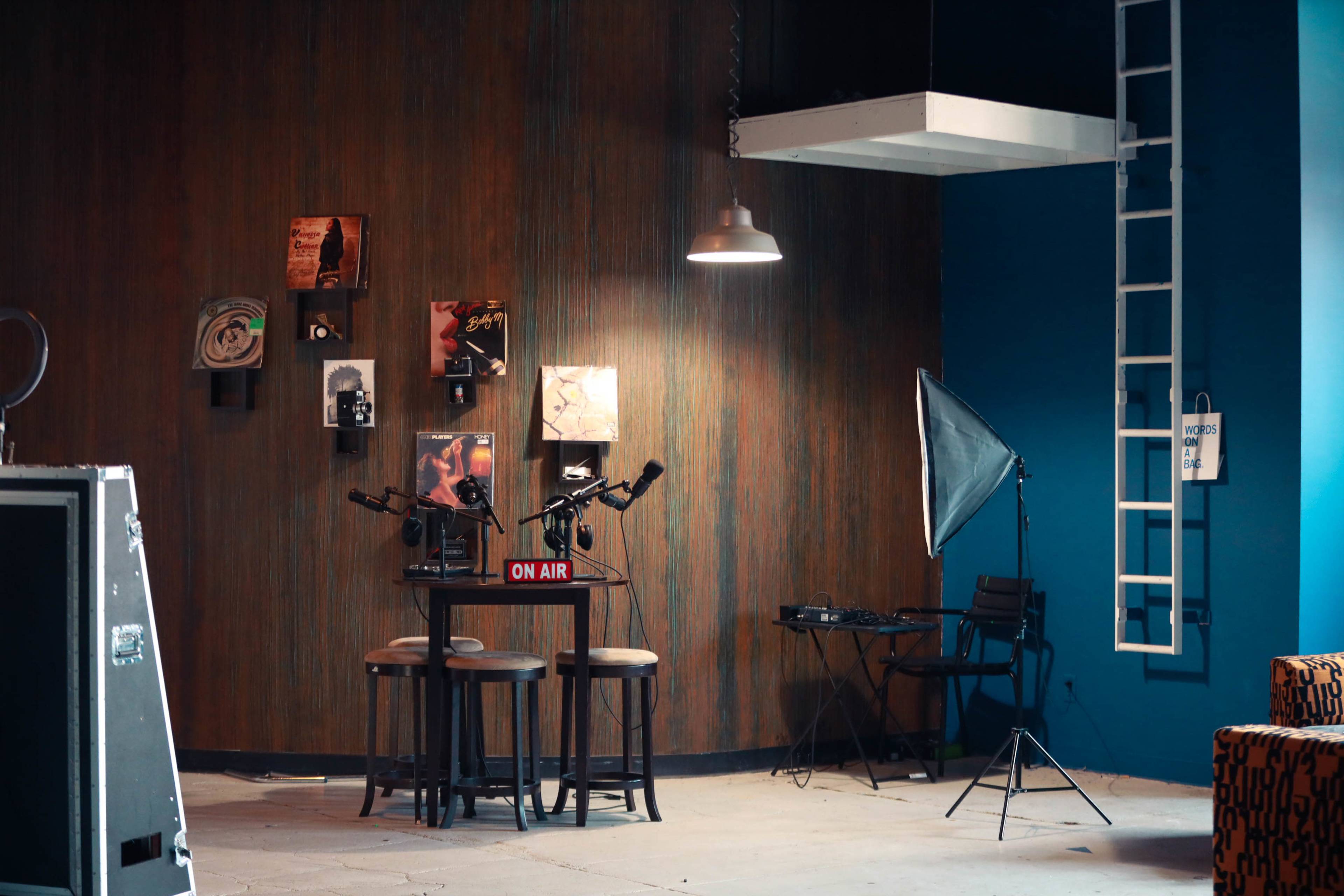 The image features a room with a wooden wall displaying framed art, two microphones on stands facing three stools, and a spotlight beside a backdrop and a folding chair.
