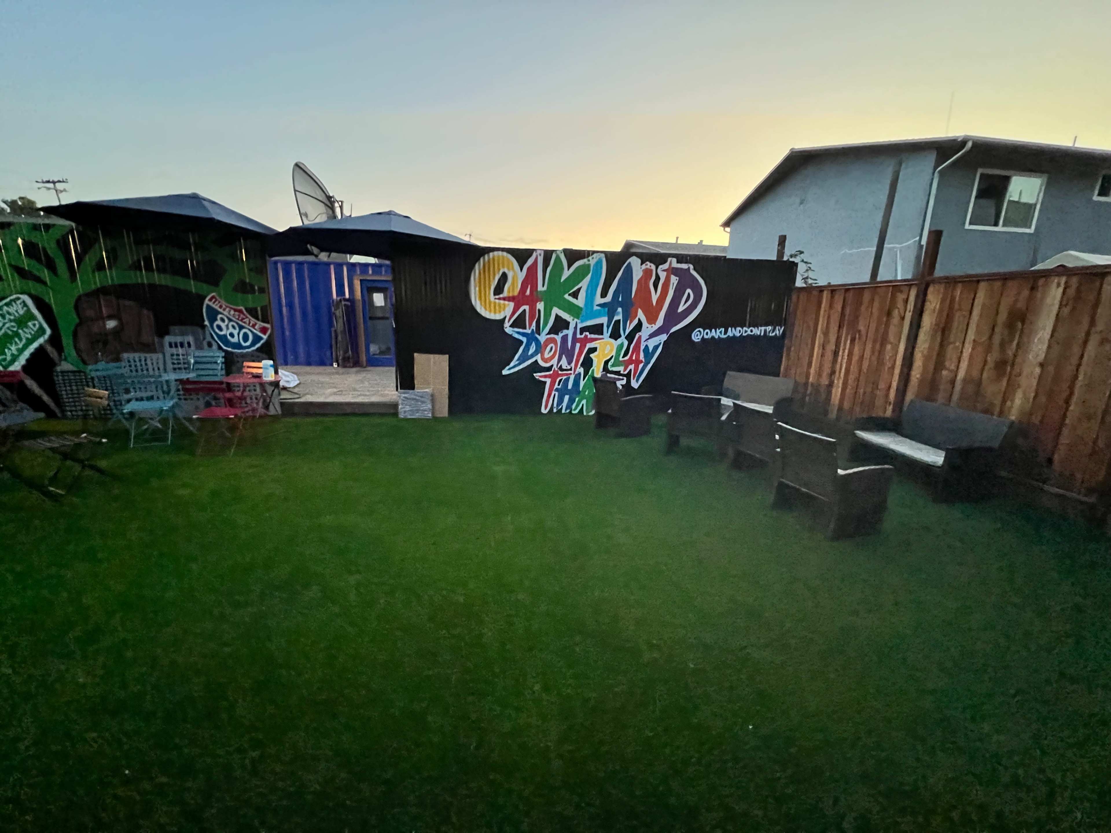 The image shows a small outdoor space with green grass, lawn chairs, and a mural on a wooden fence that reads "OAKLAND DON'T TELL."