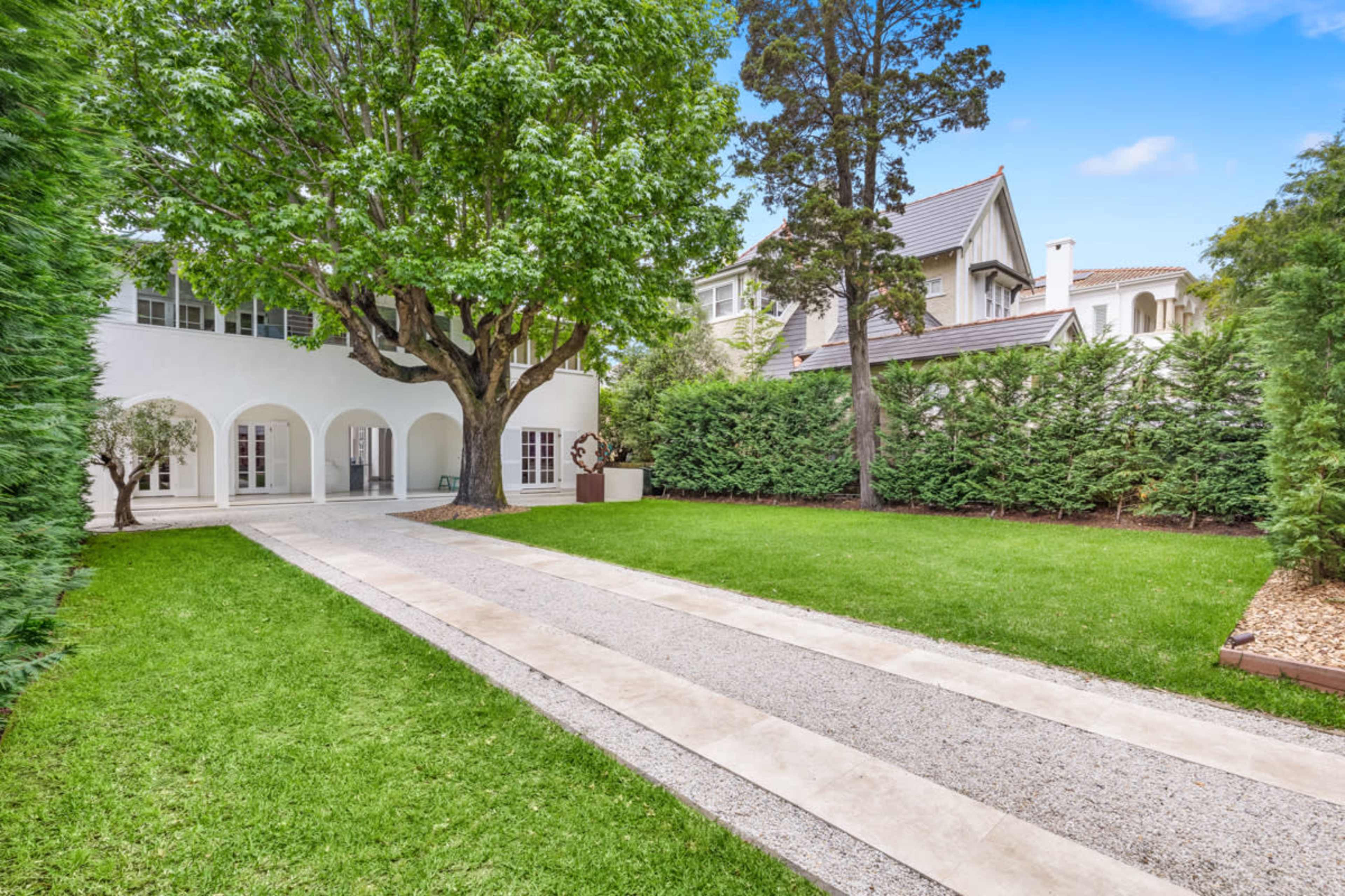 A gravel pathway leads to a white-walled house surrounded by manicured lawns and tall greenery.