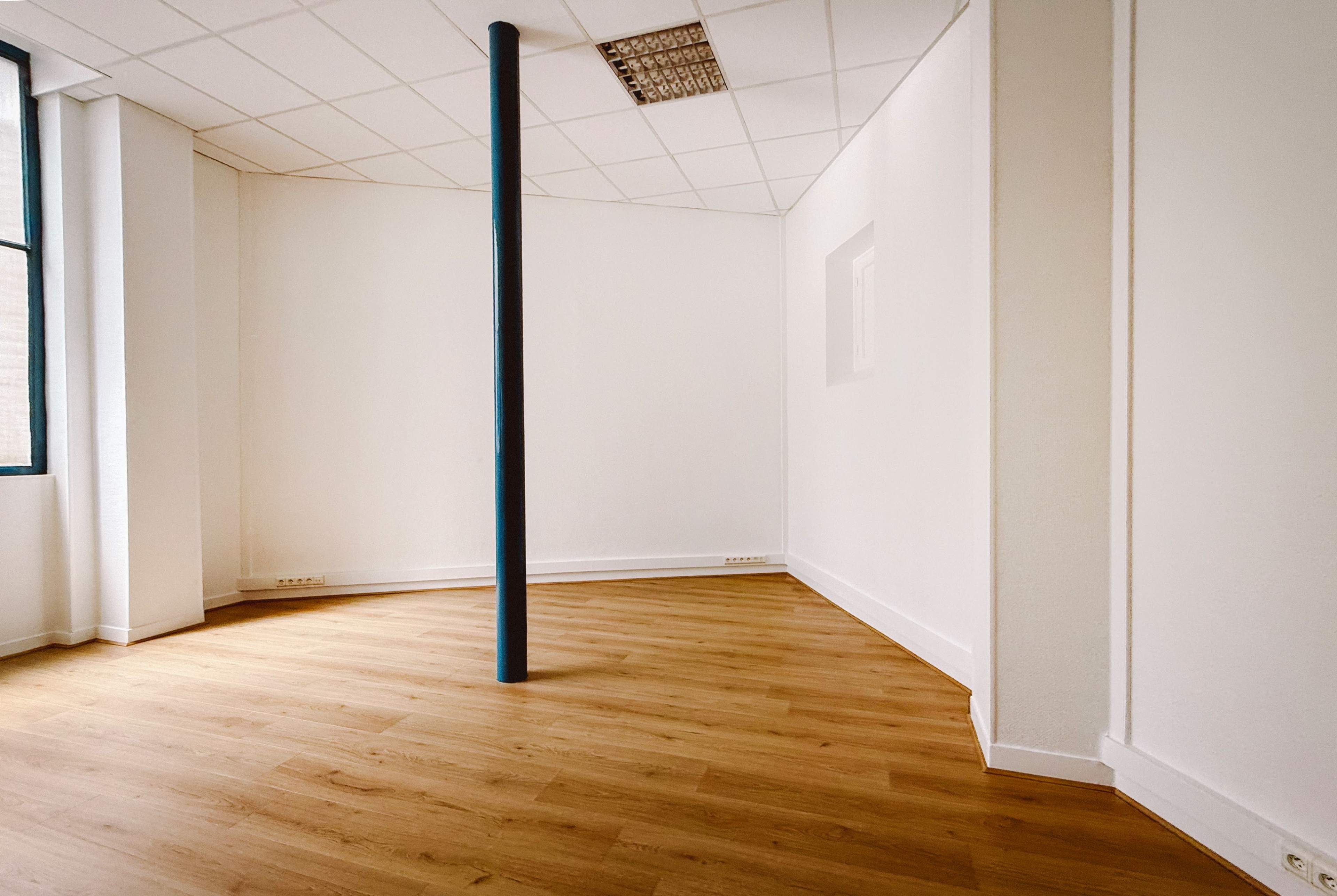 The image shows a minimally furnished corner of a room with light-colored wood flooring and a central blue support column.