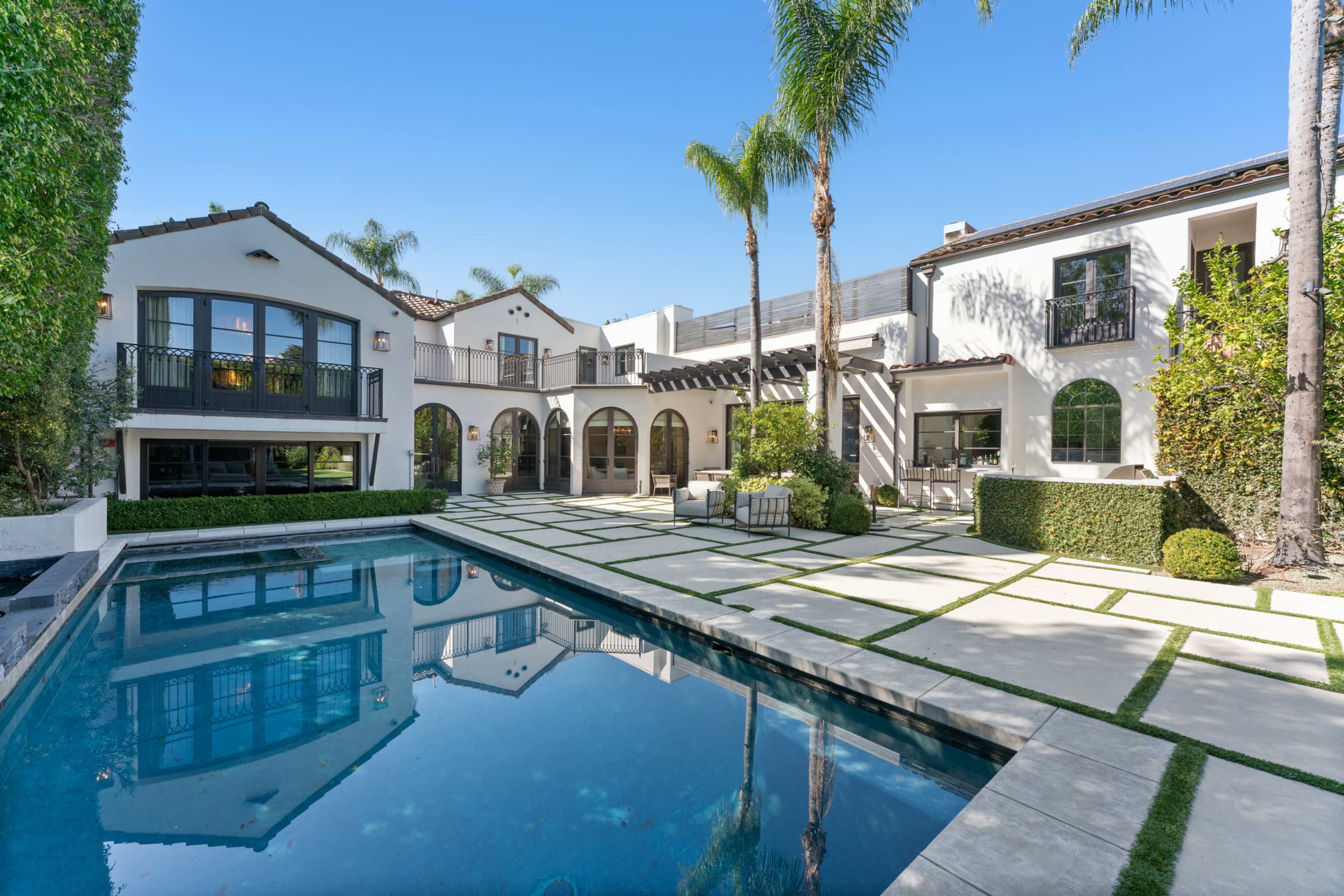 The image shows a modern Mediterranean-style home with a pool surrounded by a landscaped patio and palm trees.