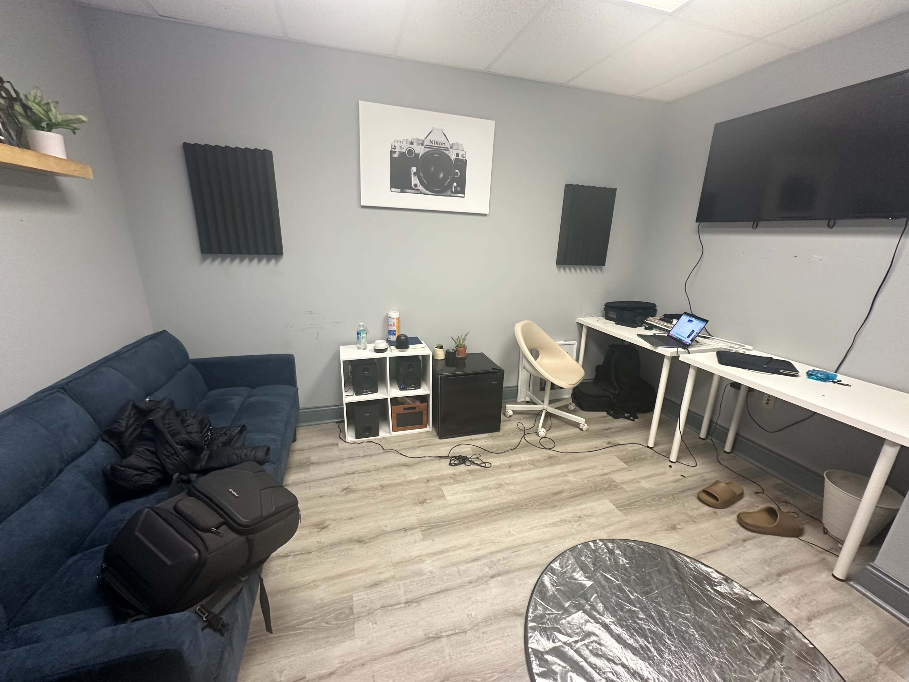 The room features a blue couch, a white desk with a computer, and organizers against a gray wall, with soundproofing panels on either side and a large TV.