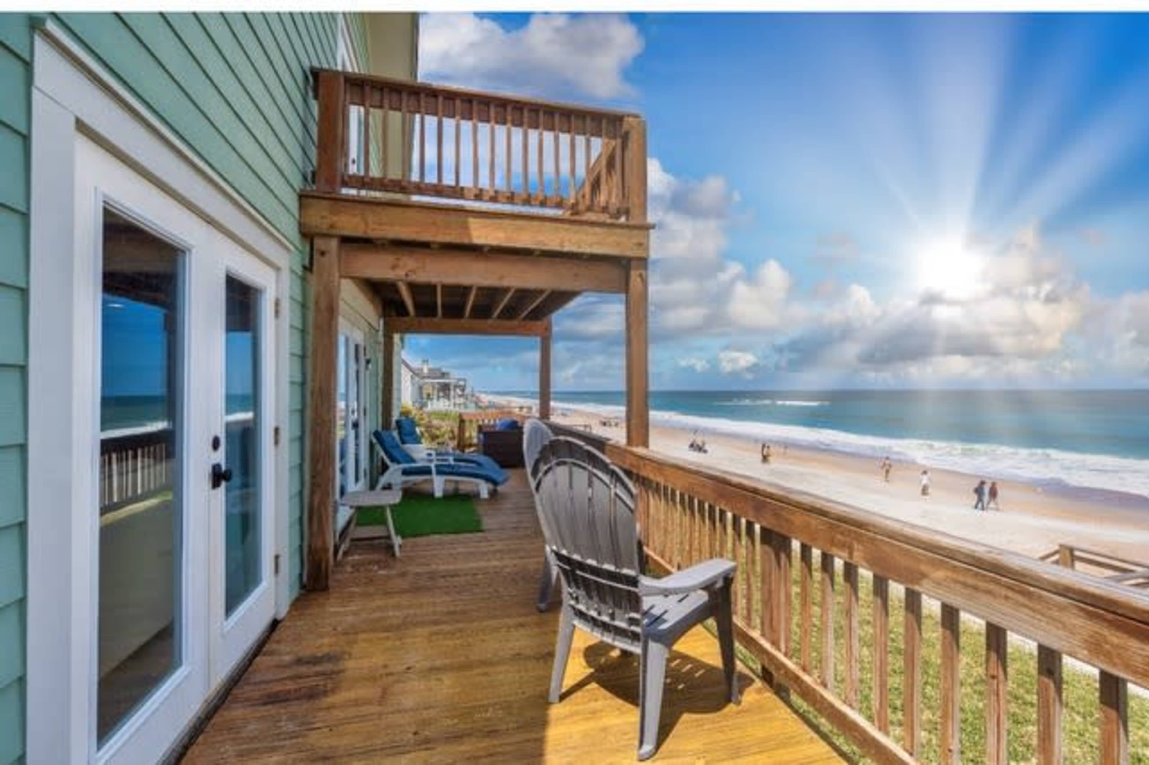A wooden deck with two chairs overlooks a sandy beach where people stroll along the shore under a bright sky.