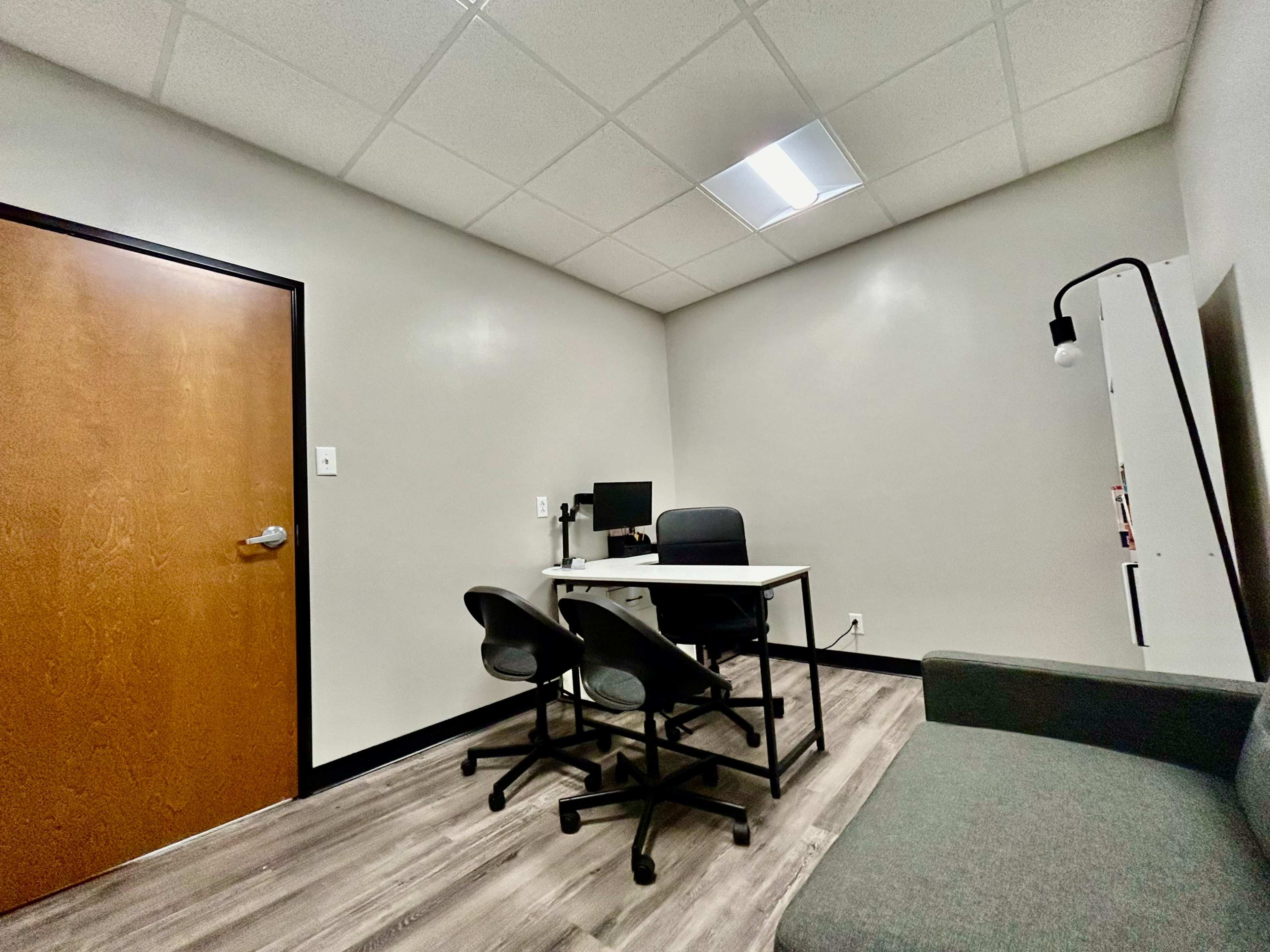 A sparsely furnished office space with a desk, two chairs, a computer, and a couch against the wall.