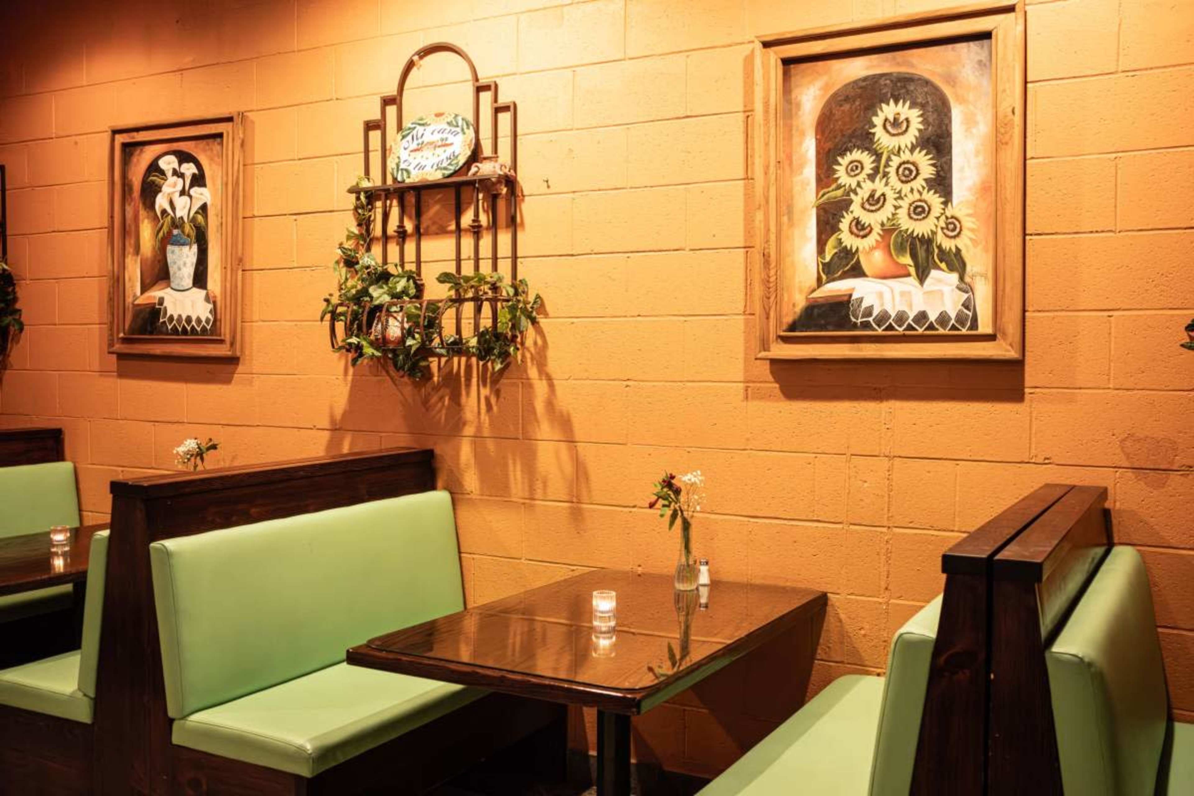The image shows a cozy restaurant interior with green booth seating, a wooden table, and wall art featuring floral paintings.