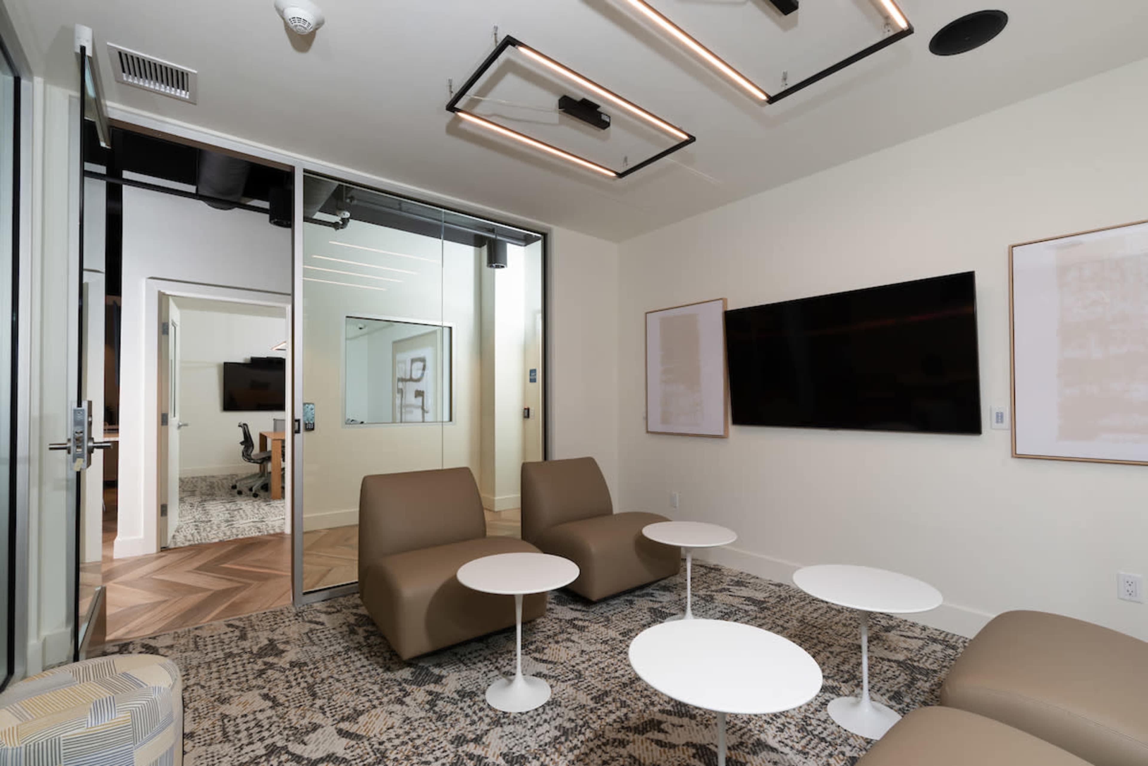 A modern meeting room with two brown lounge chairs, a set of small white tables, and a wall-mounted TV.