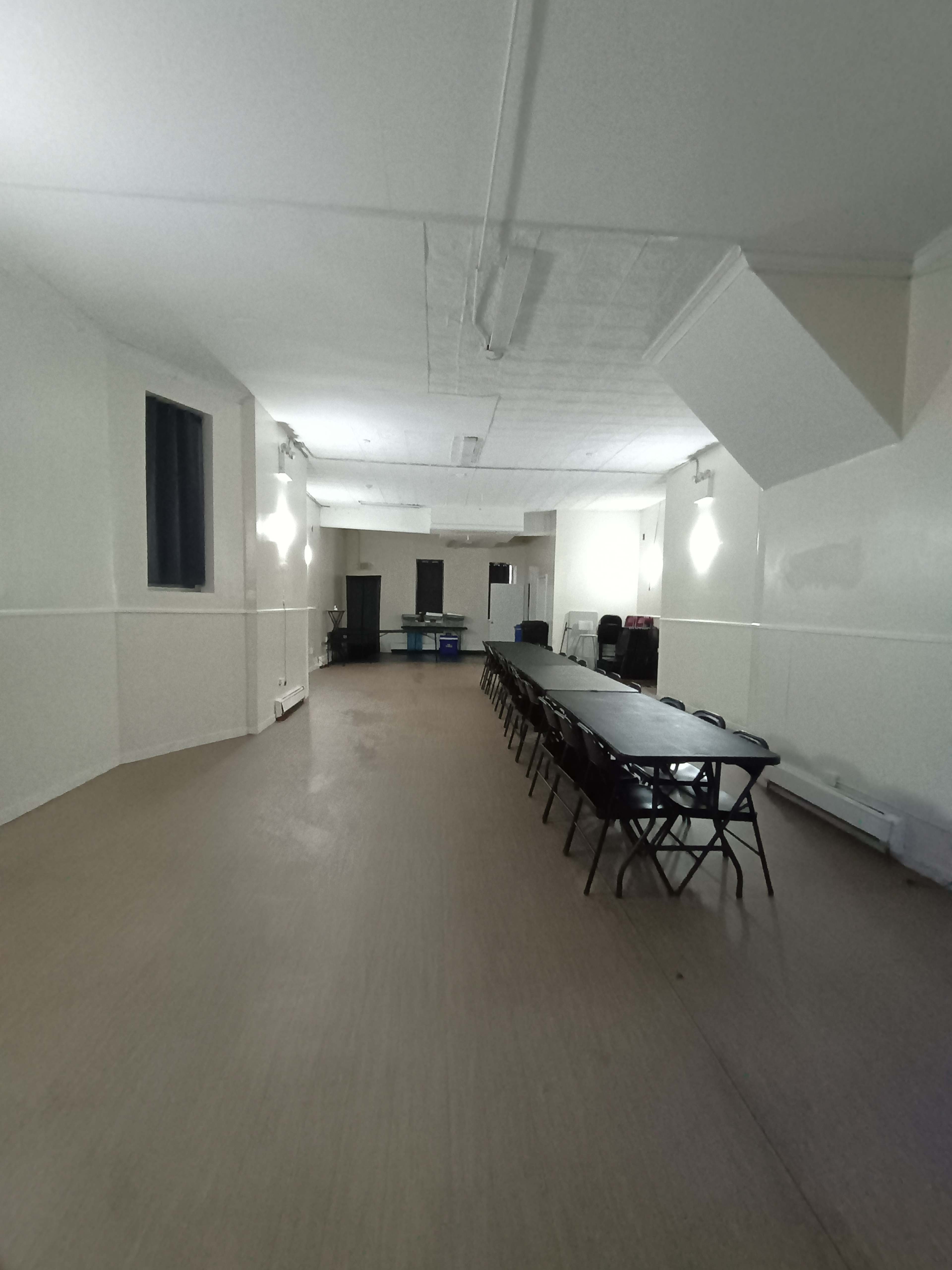 The image shows a large, empty room with a long row of tables and chairs set up along the center, illuminated by overhead lights.