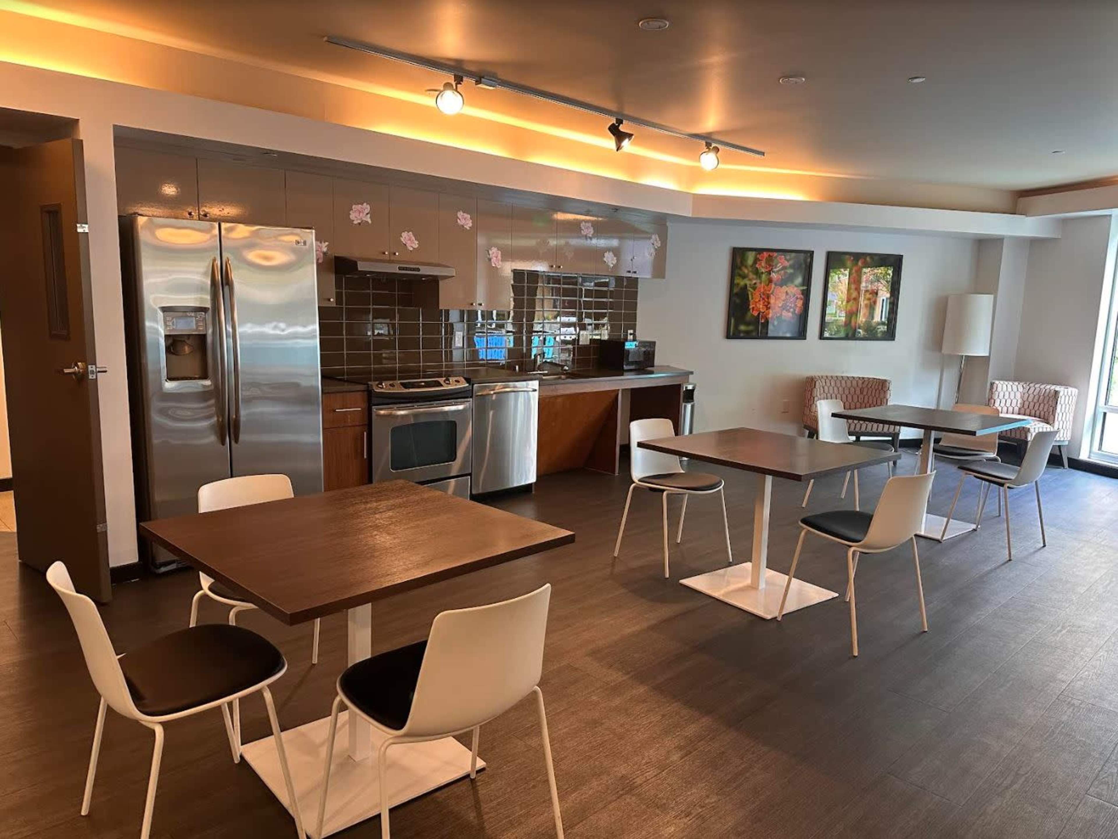 A modern kitchenette area features stainless steel appliances, wooden tables, and white chairs arranged around the space.