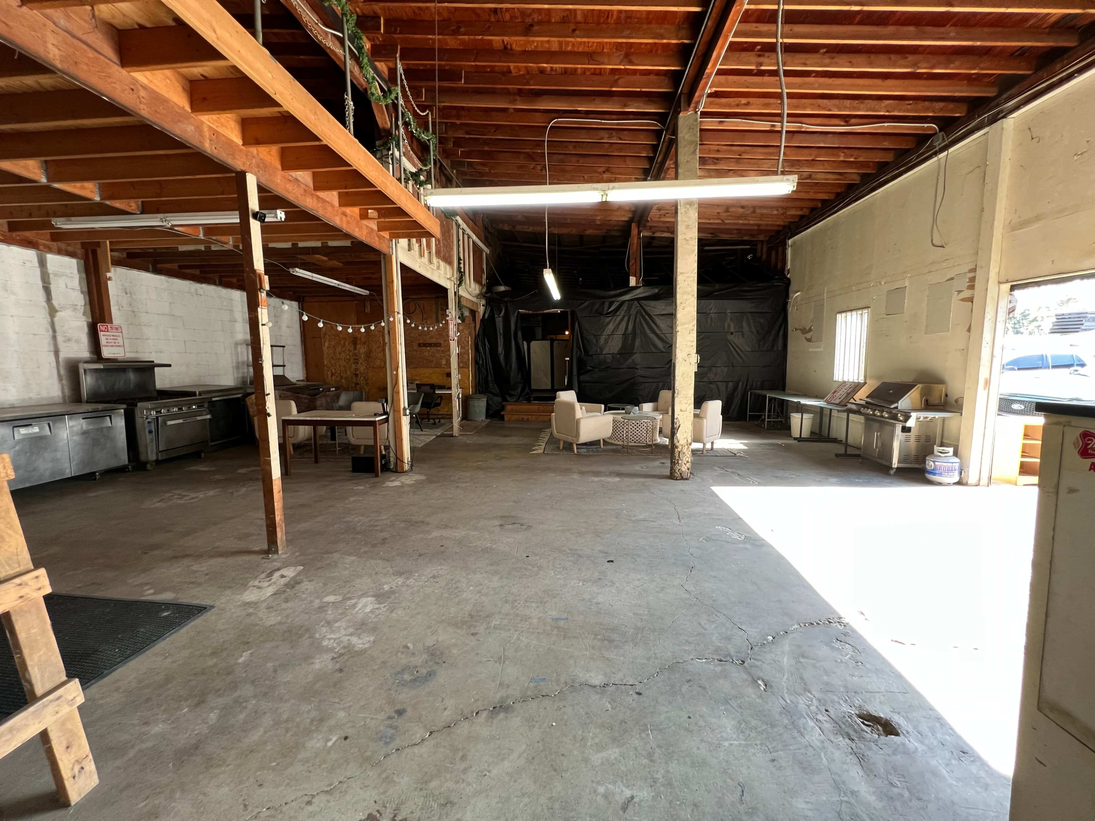 A large, open indoor space with exposed wooden beams, a commercial kitchen area on one side, and a seating area with chairs on the other, partially lit by natural light.
