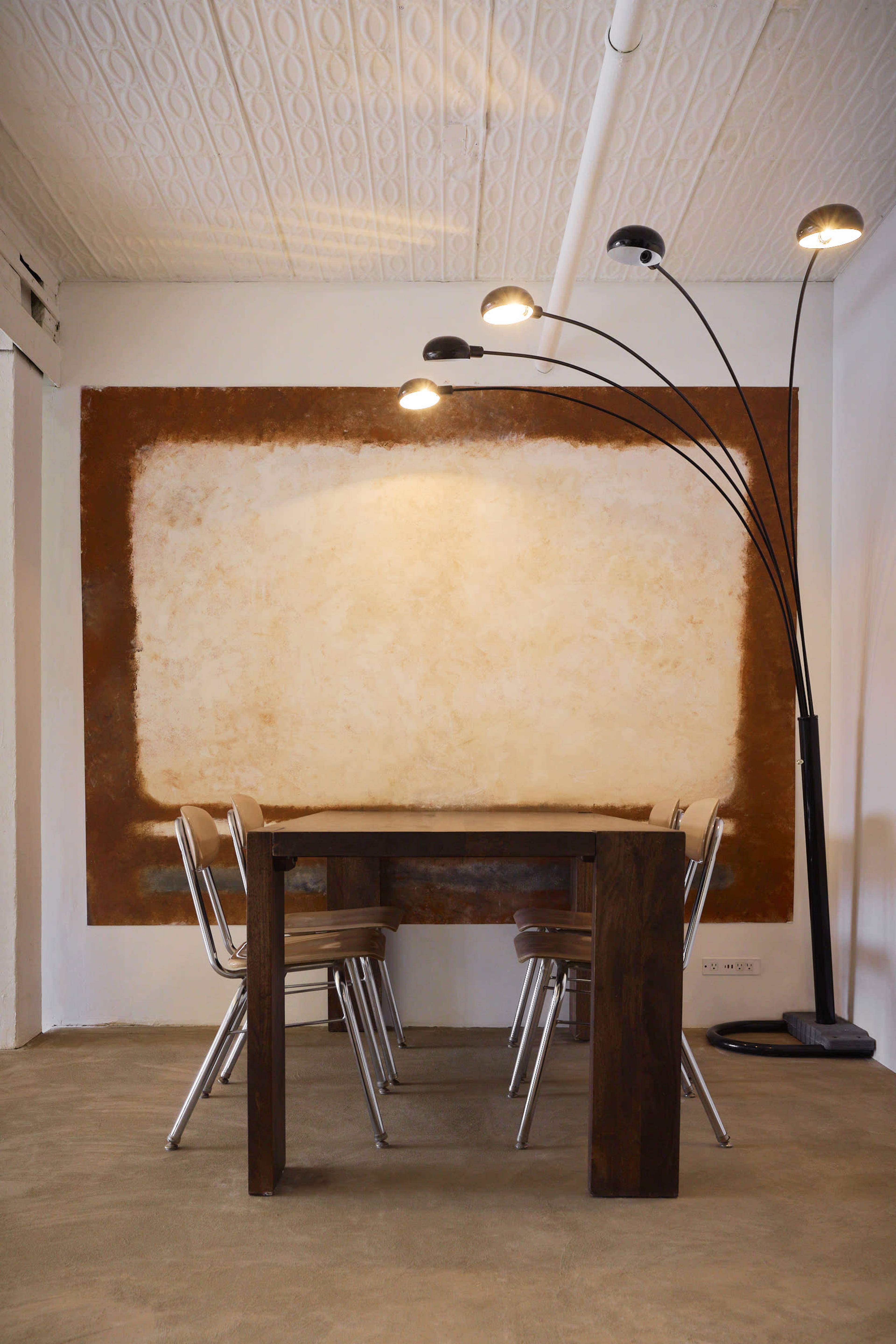 A wooden dining table with four metal chairs is positioned under an arc floor lamp in a room featuring a textured, light brown wall.