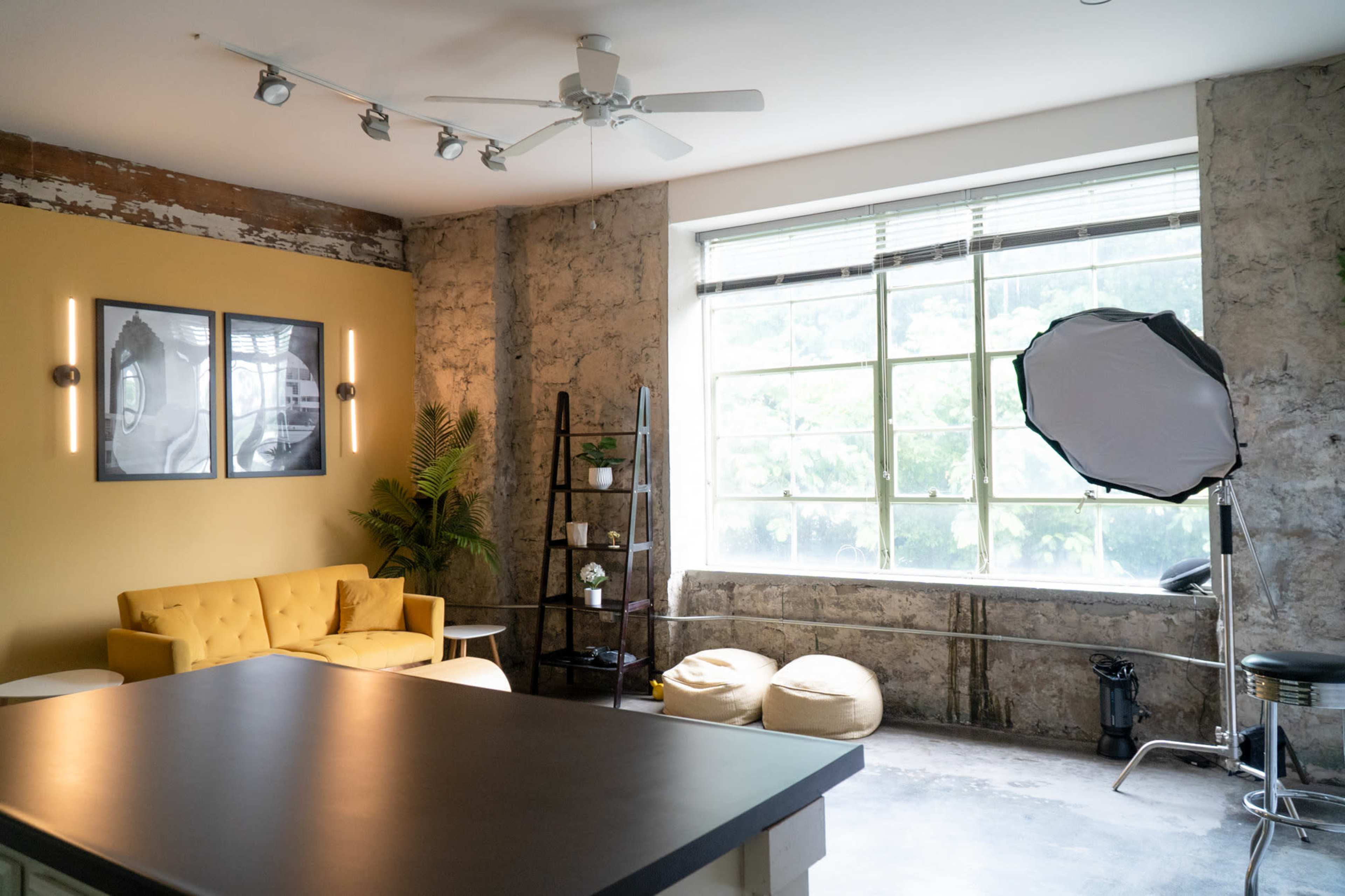 The image shows a modern indoor space with a yellow couch, large windows, and a photography setup featuring a softbox.