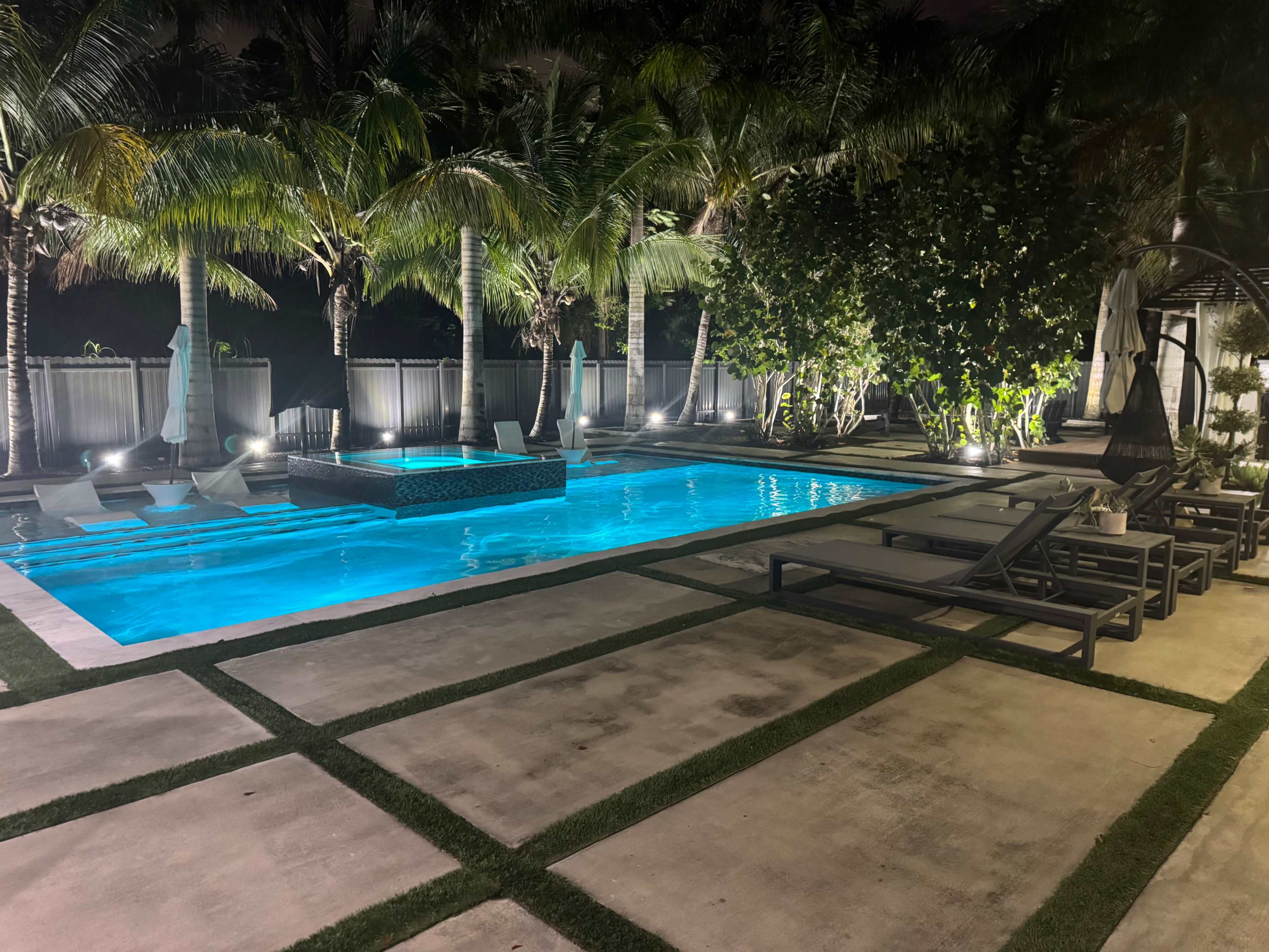 The image shows a well-lit swimming pool surrounded by palm trees and lounge chairs, with a small elevated section in the center.