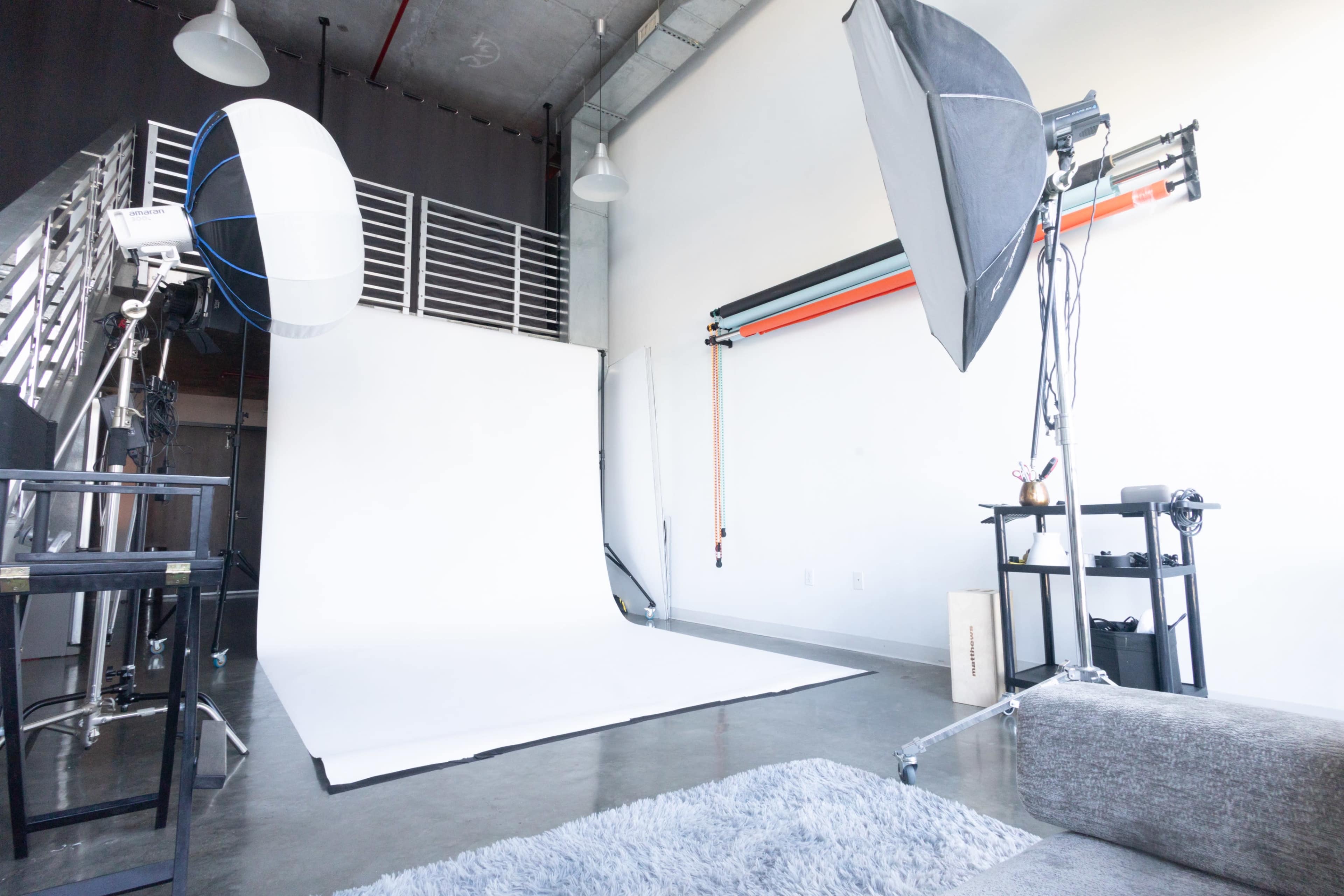 The image shows a photography studio equipped with a white backdrop, lighting equipment, and a gray rug on the floor.