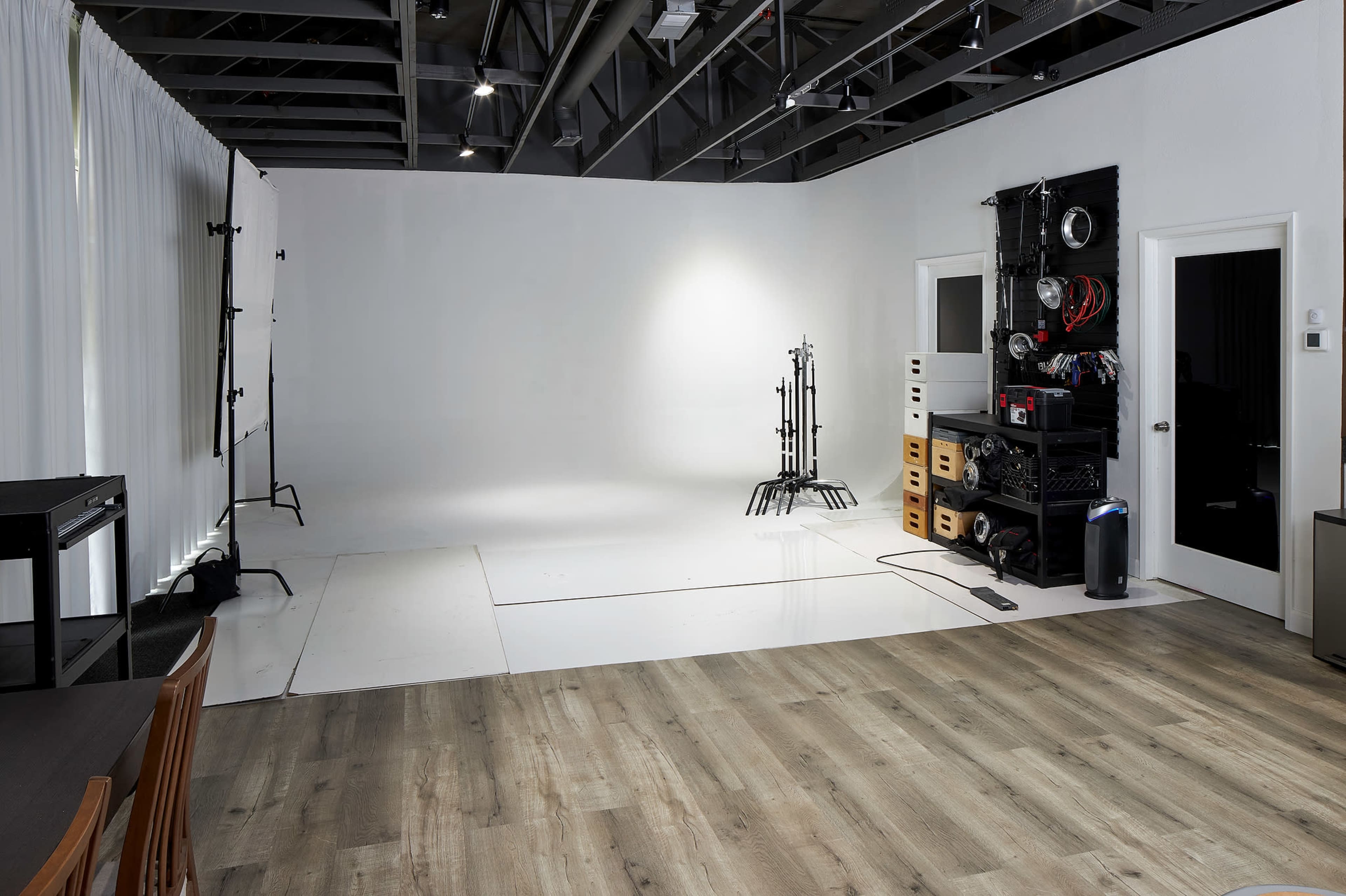 The image shows a photography studio with a white backdrop, light stands, and storage units against the walls, along with wooden flooring.