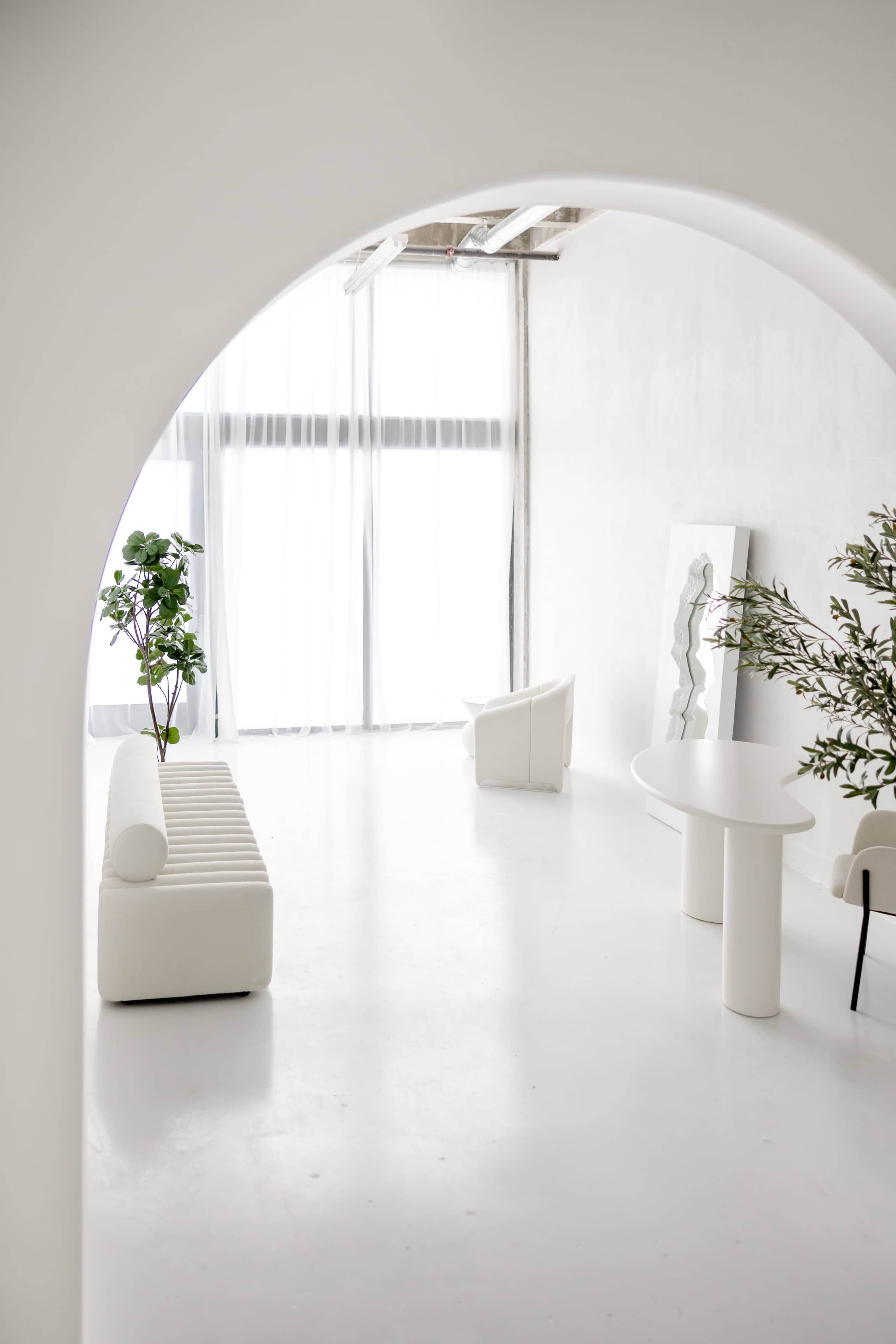 The image shows a minimalistic interior space with white walls, a sofa, a side chair, a round table, and plants near large windows allowing natural light.