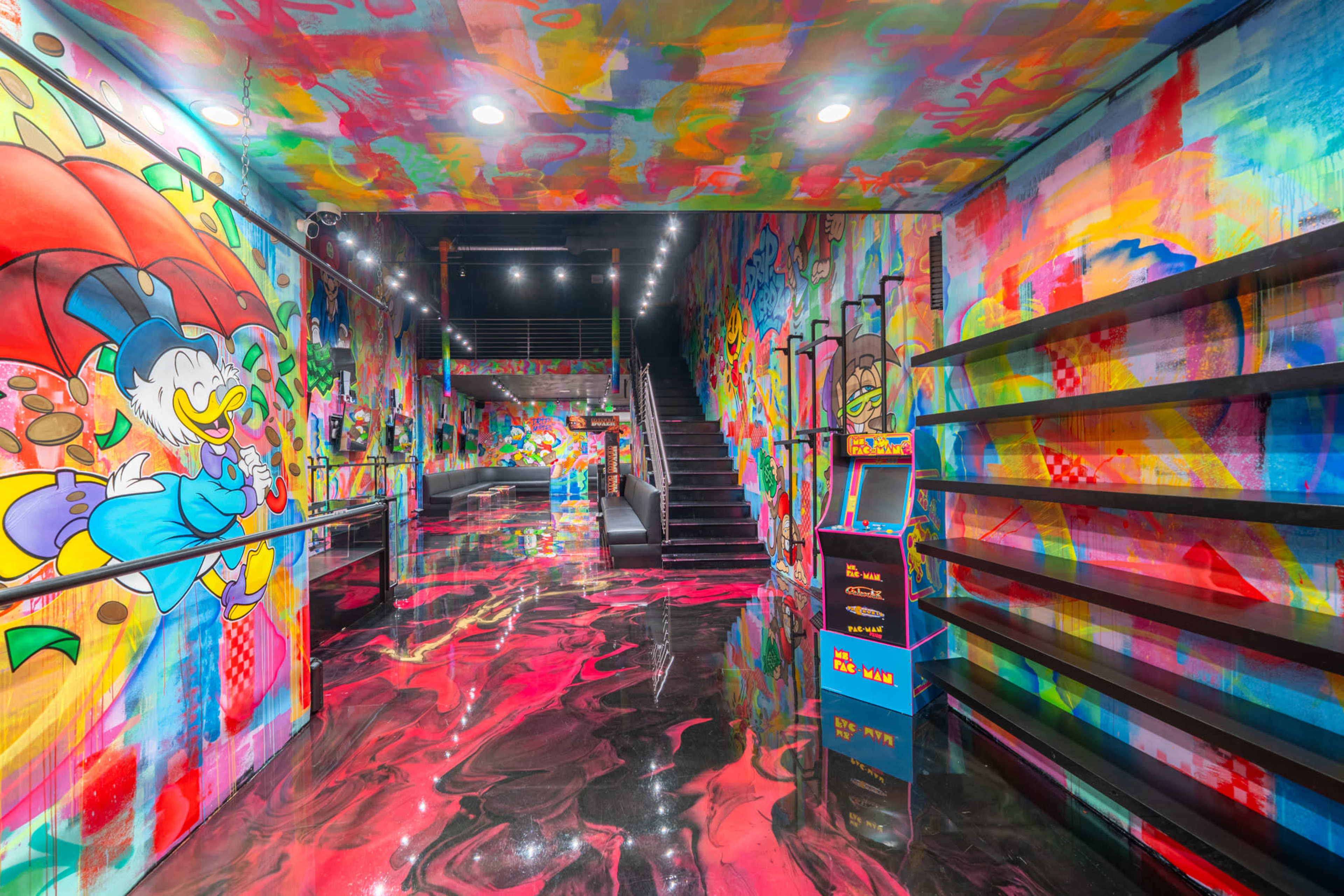 The image shows a brightly colored hallway with vibrant graffiti-style murals on the walls and a glossy, multicolored floor, leading to a staircase.