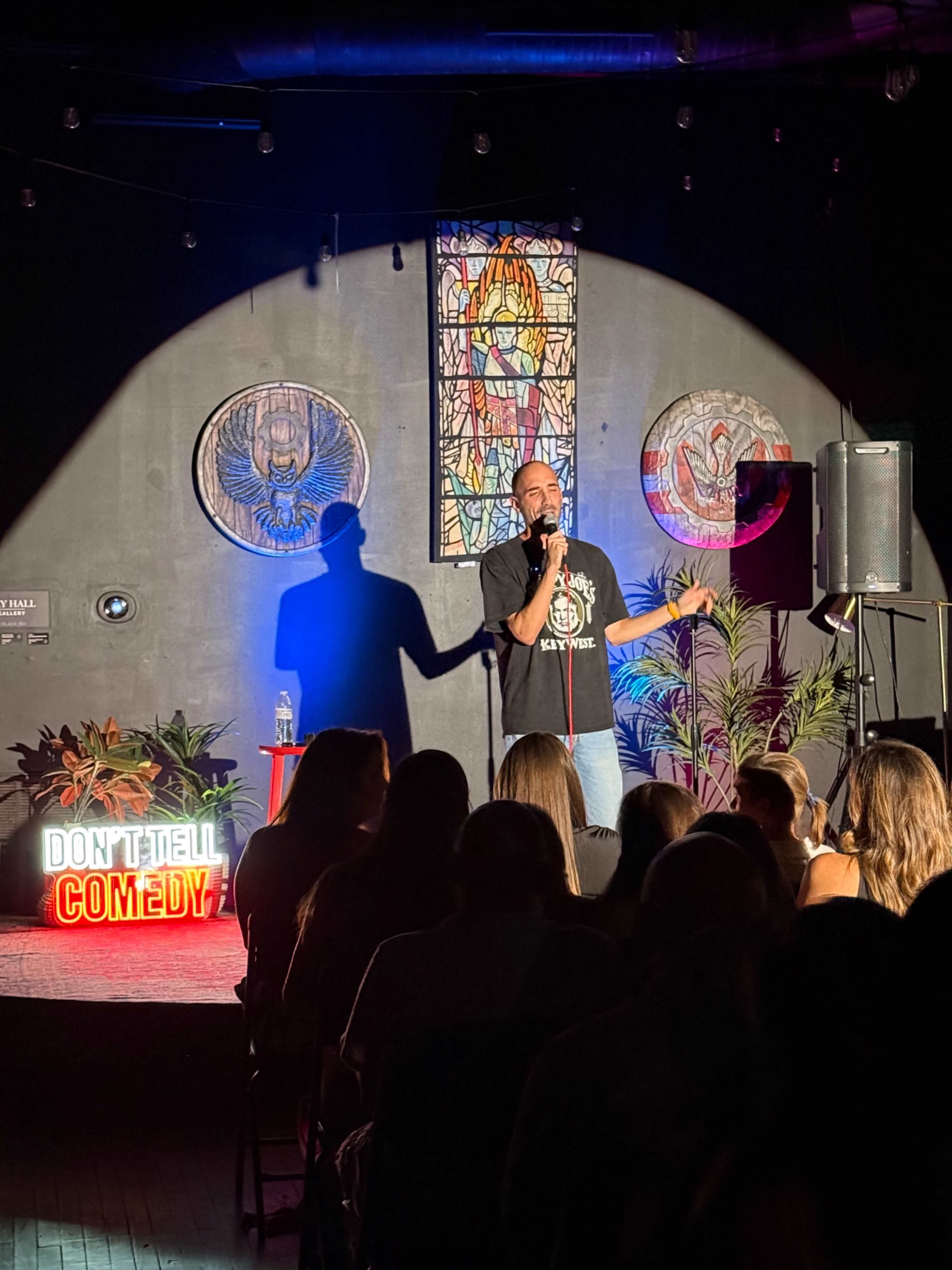 A comedian performs on stage in front of an audience, with colorful artwork and a neon sign reading "DON'T TELL COMEDY" in the foreground.