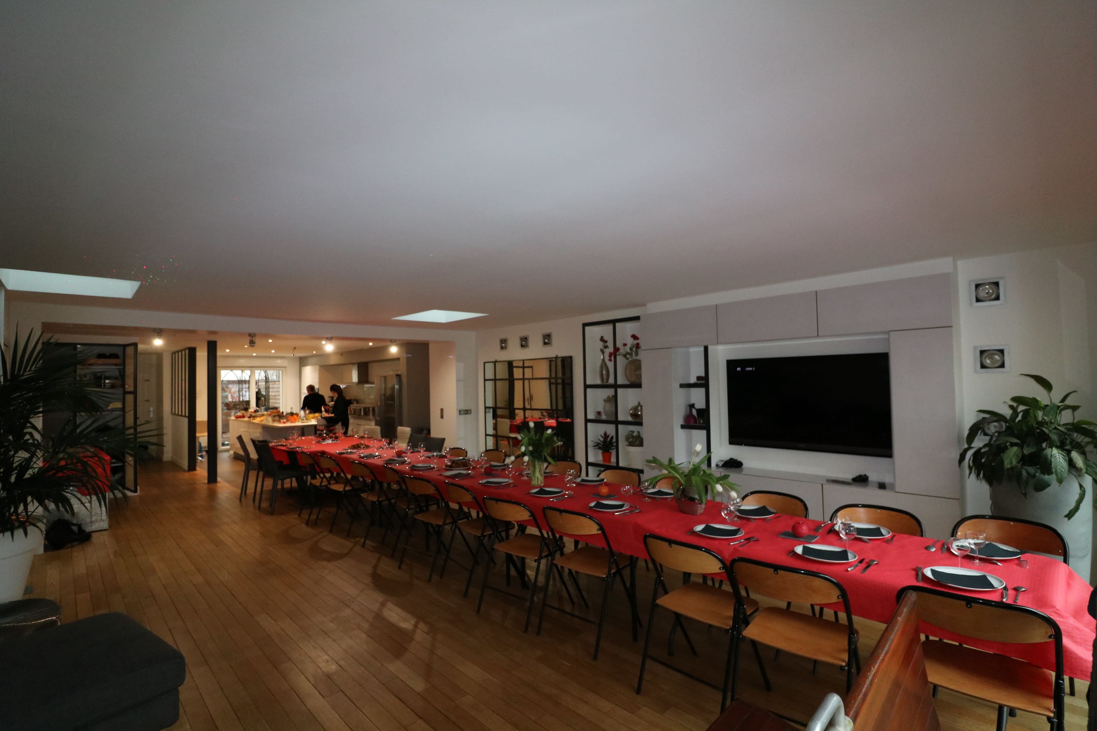 A large dining area features a long table set with red tablecloths and dinnerware, while the background shows a kitchen and seating area.