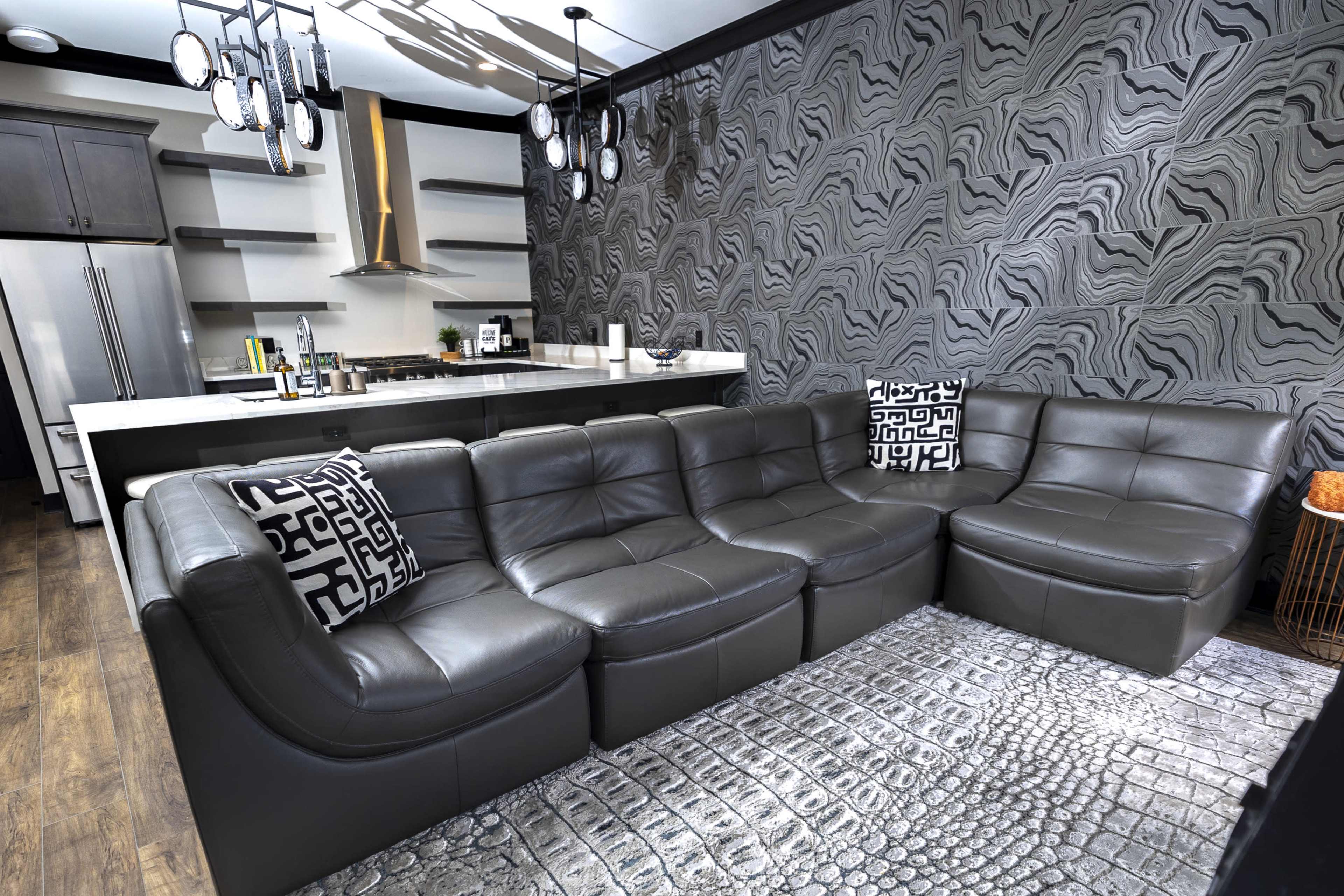 The image shows a modern living room with a dark gray leather sectional sofa, patterned throw pillows, and a patterned rug, alongside a sleek kitchen with a stainless steel oven and minimalist cabinetry.