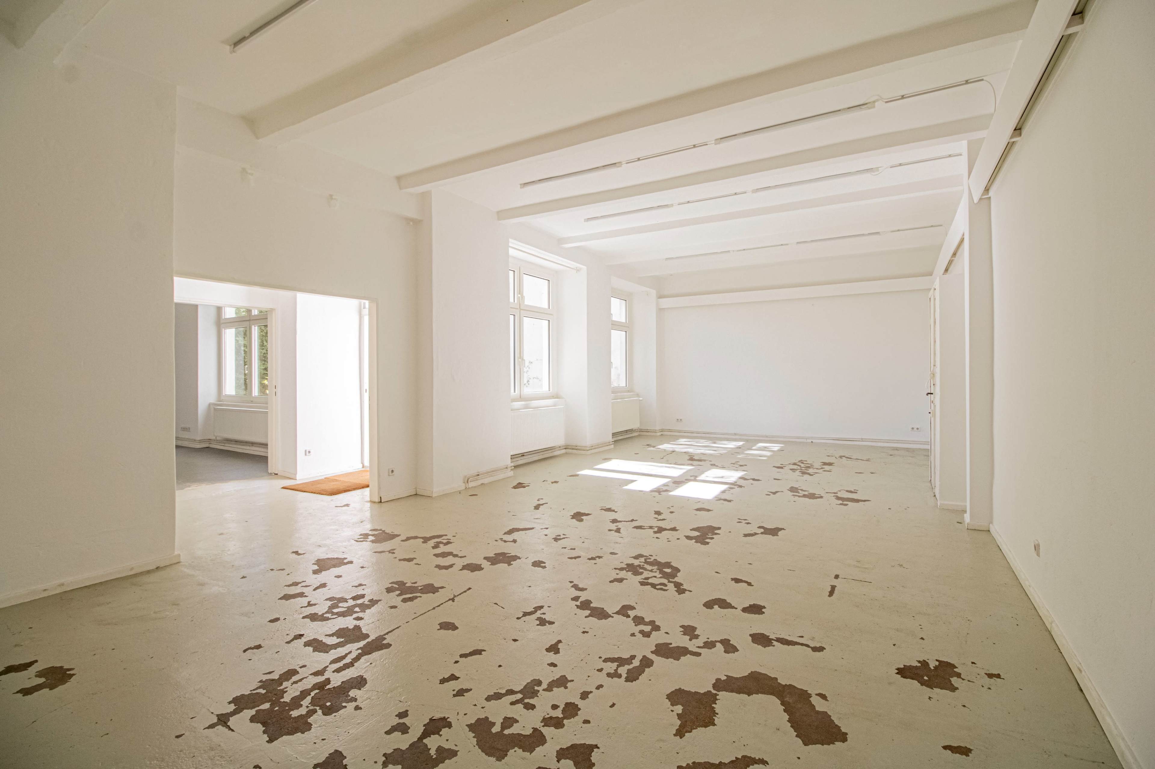 The image shows a spacious, empty room with white walls and a partially worn floor, featuring large windows that allow natural light to enter.