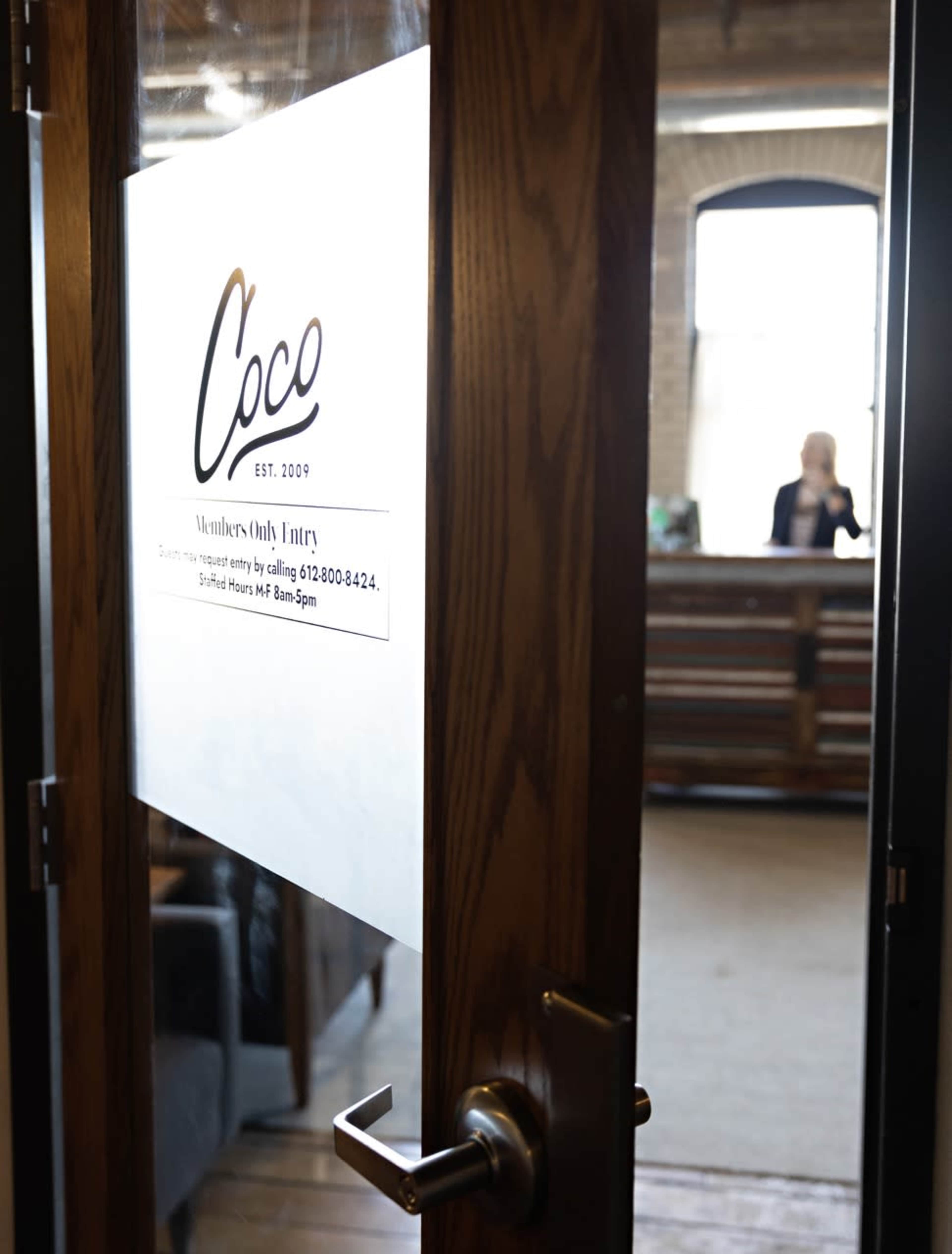 A glass door with a signage for Coco, indicating members-only entry, partially opens to reveal a woman working at a reception desk in a well-lit interior.