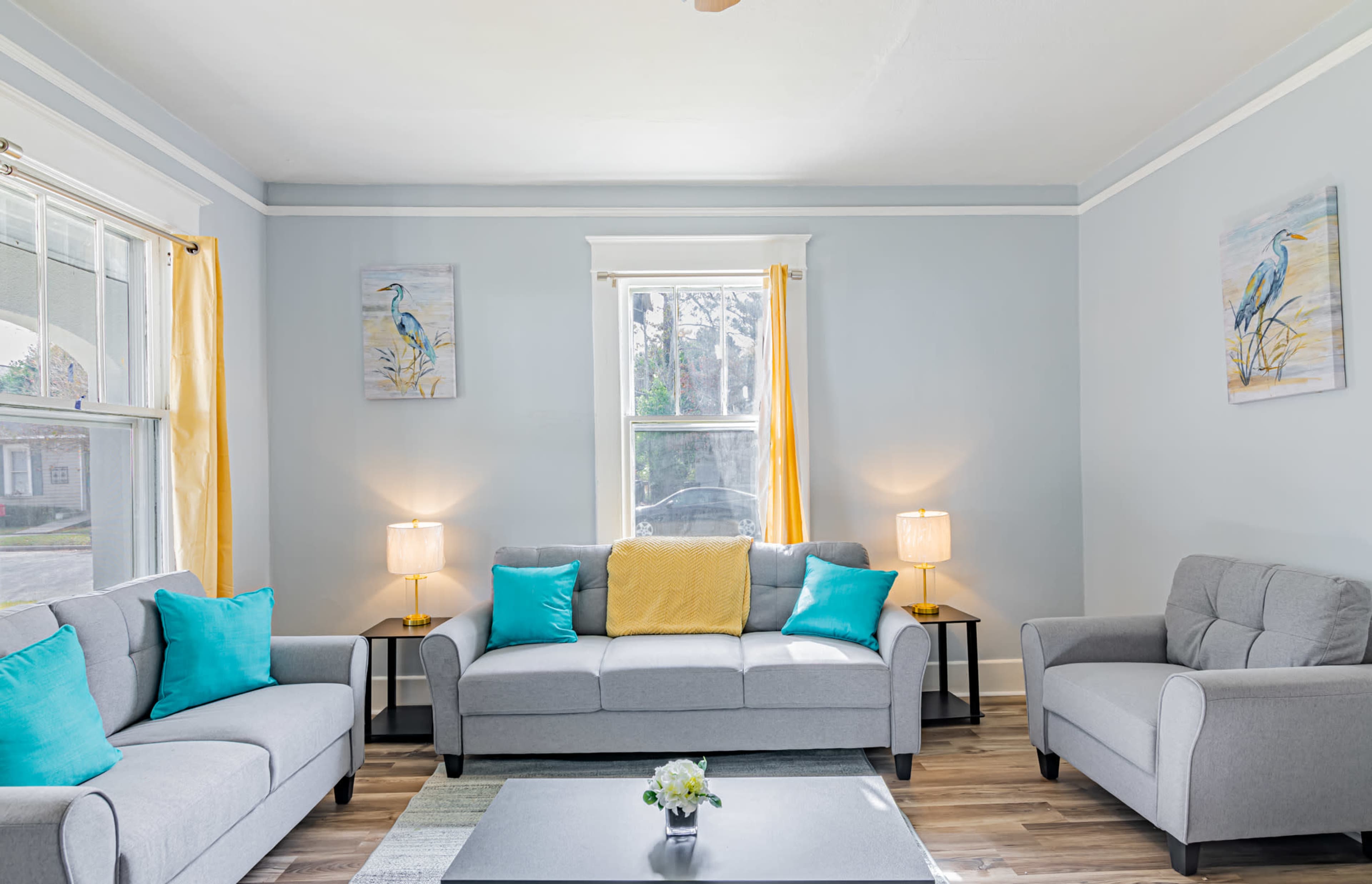 The living room features a gray sofa set with blue and yellow accent cushions, two lamps on side tables, and light-colored walls.