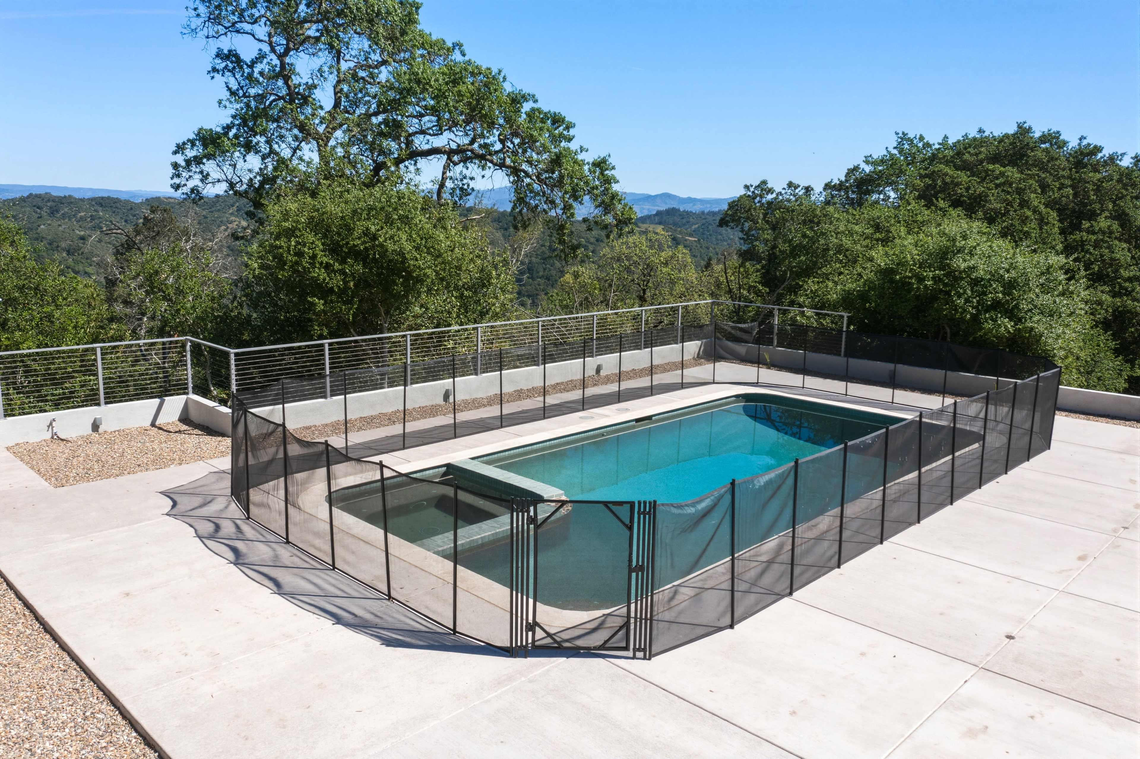 The image shows an empty swimming pool surrounded by a safety fence, situated on a concrete deck with trees in the background.