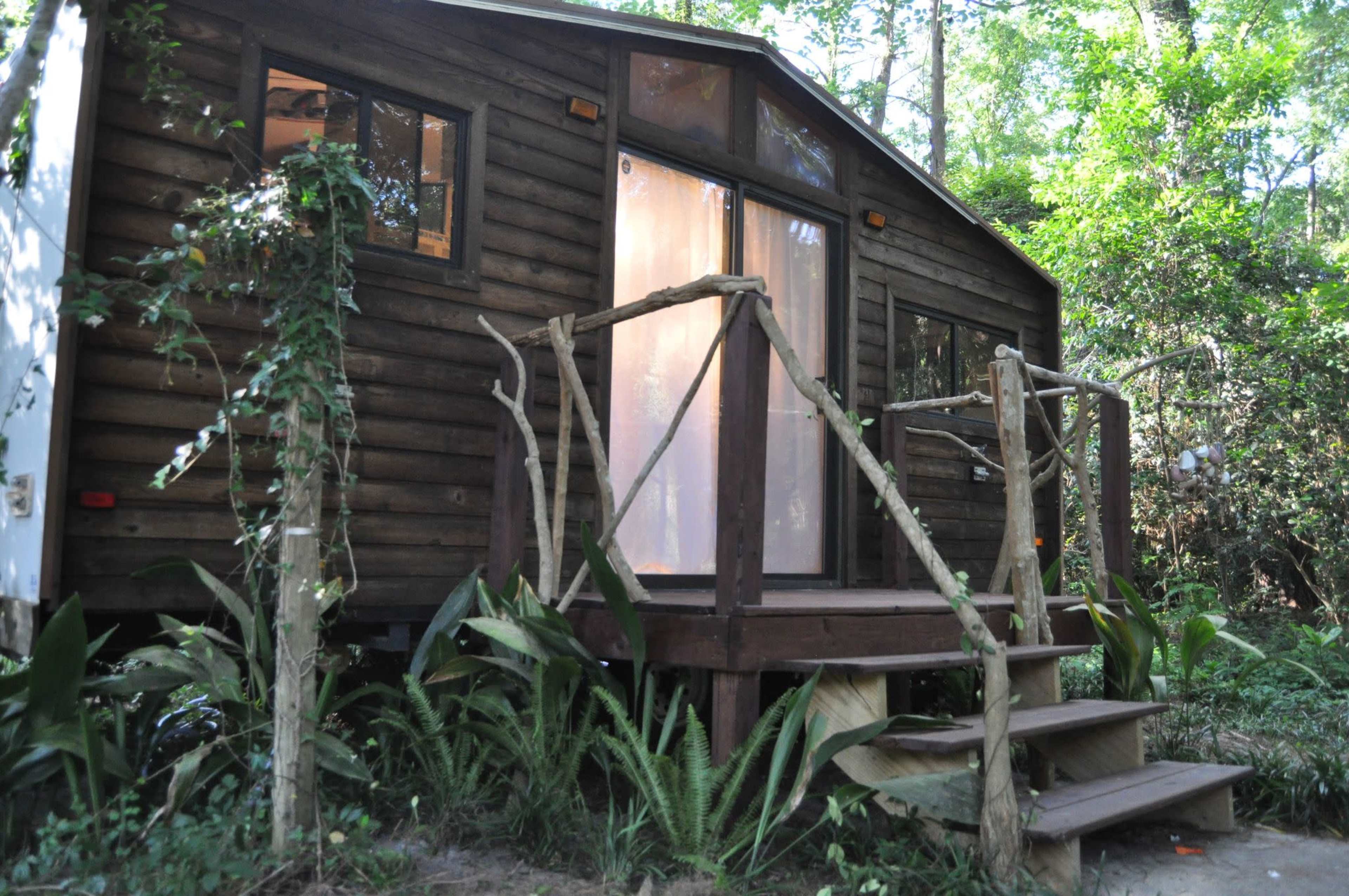 The image shows a wooden cabin with a deck and steps, surrounded by greenery.