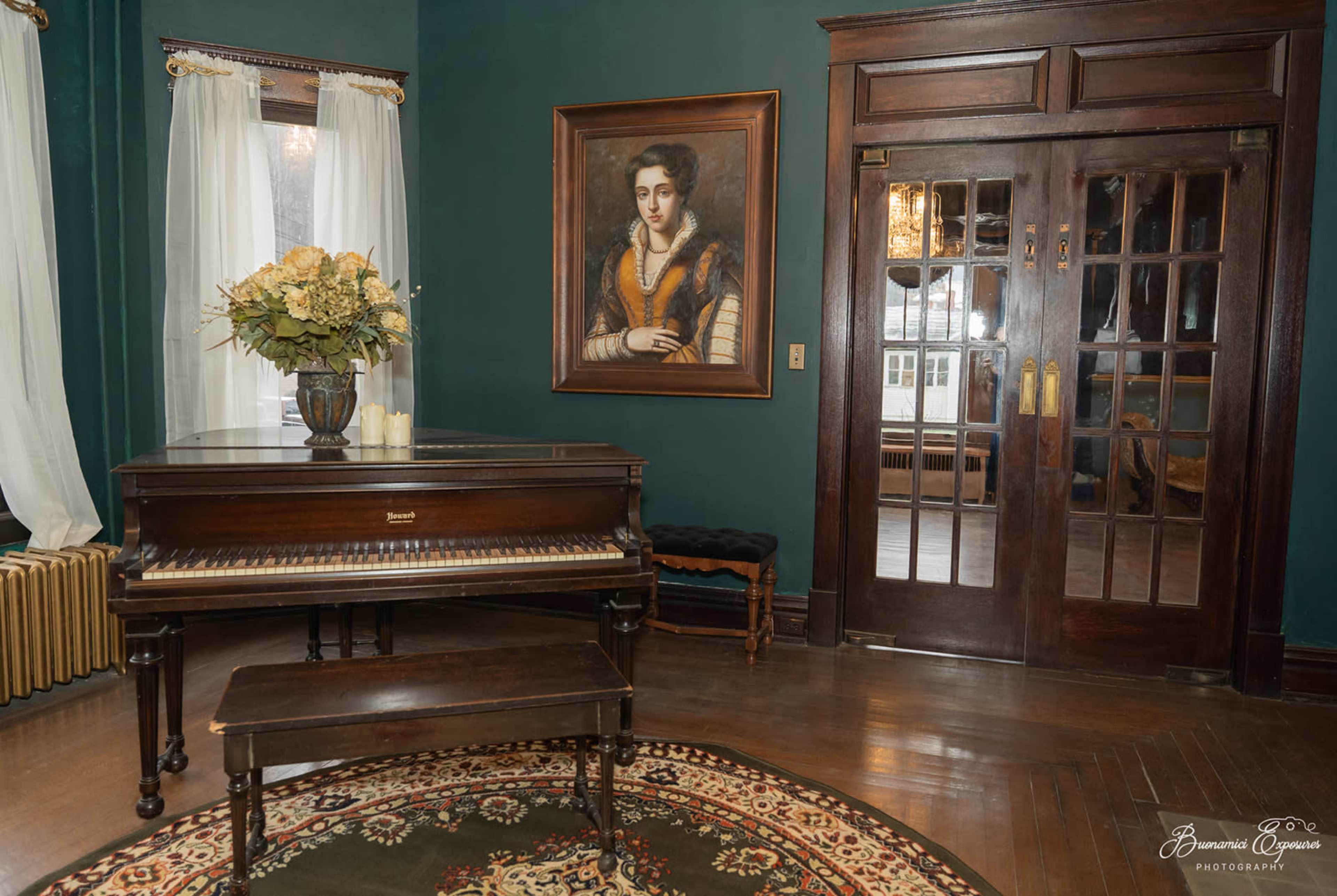 A grand piano sits in a room with dark green walls, a large floral arrangement on top, and a portrait hanging nearby.
