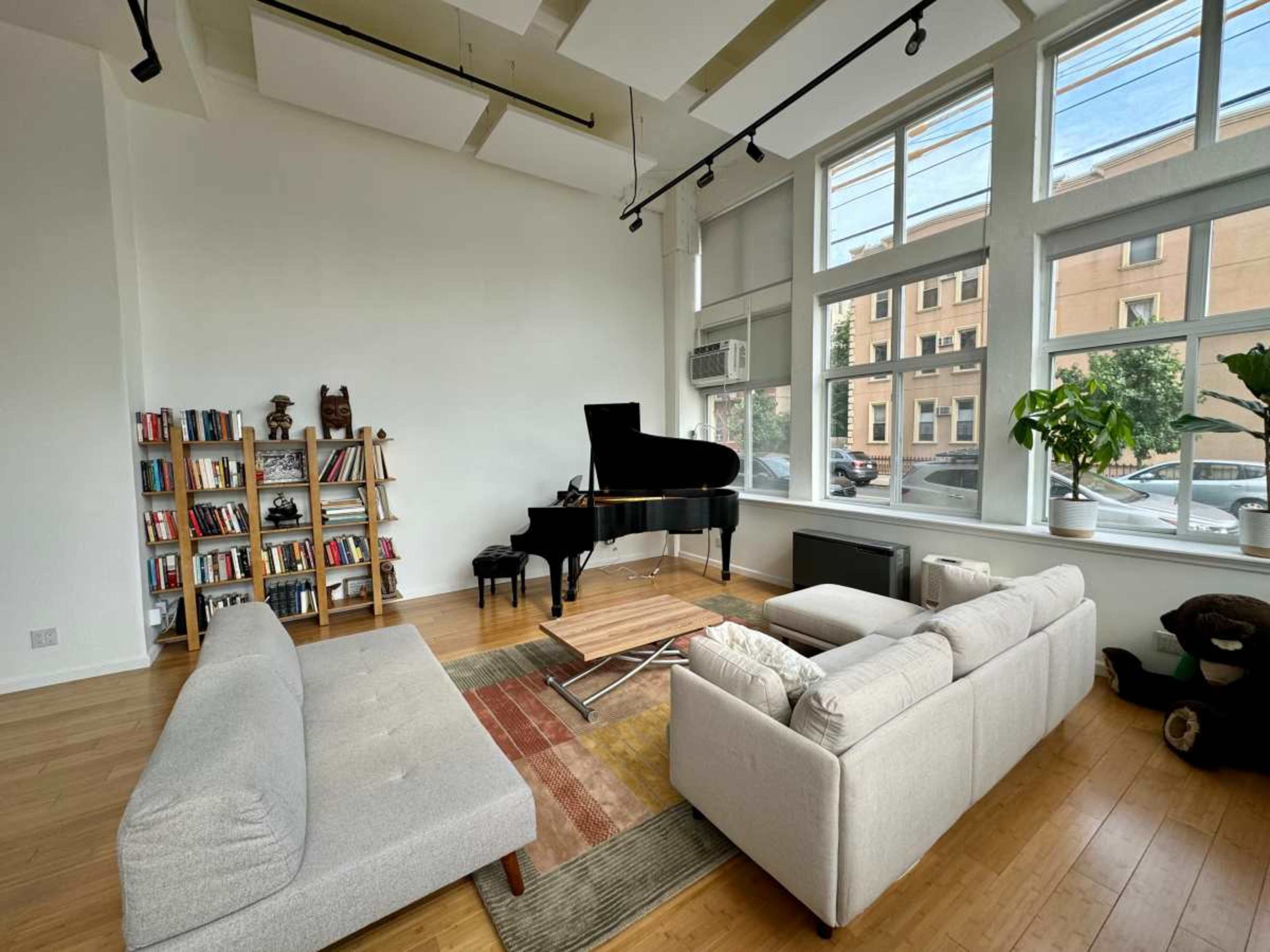 The spacious room features a grand piano, a bookshelf filled with books, a light-colored sofa, and large windows letting in natural light.