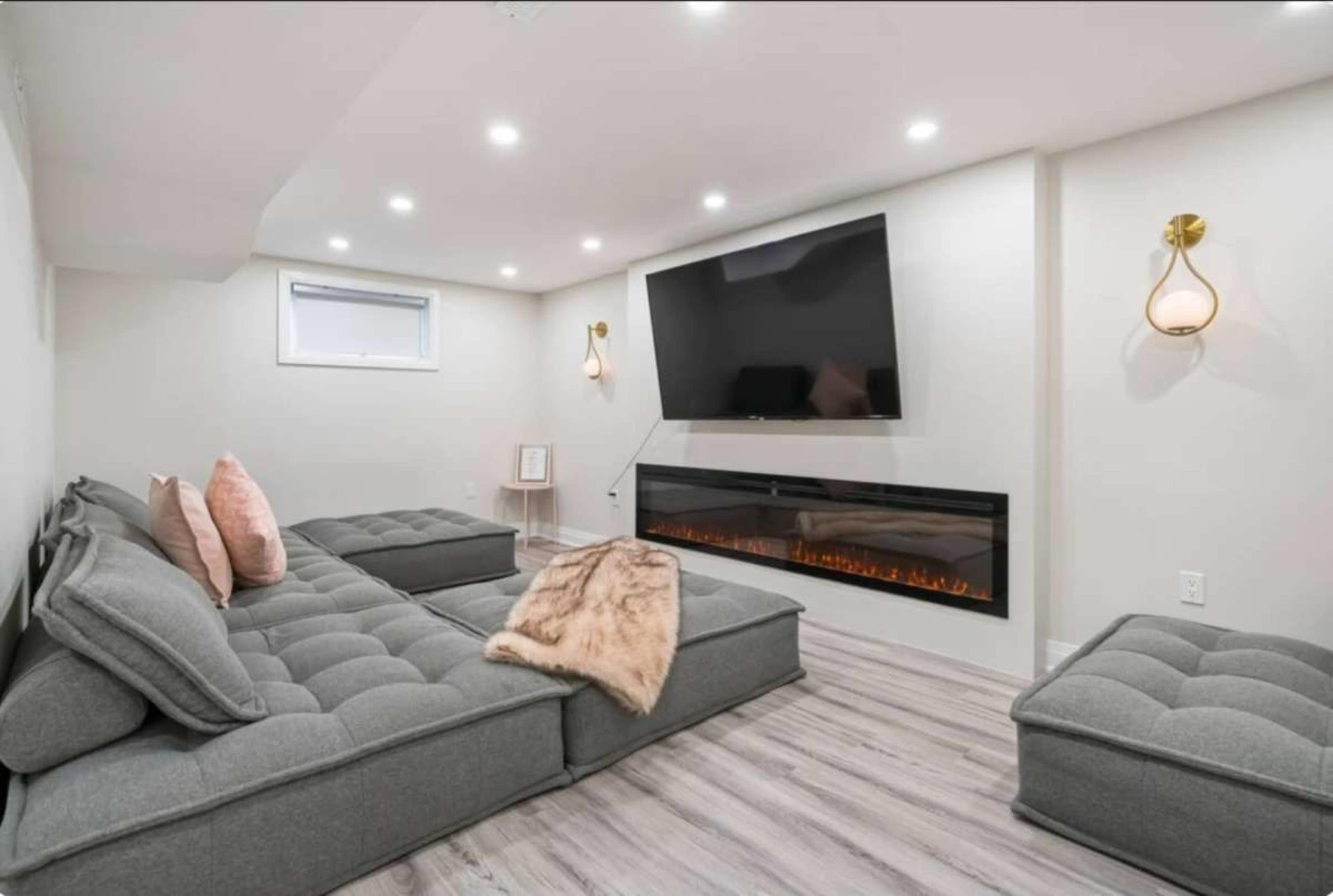 The image shows a modern living room with a large flat-screen TV mounted on the wall, a gray sectional sofa, and an electric fireplace beneath it.